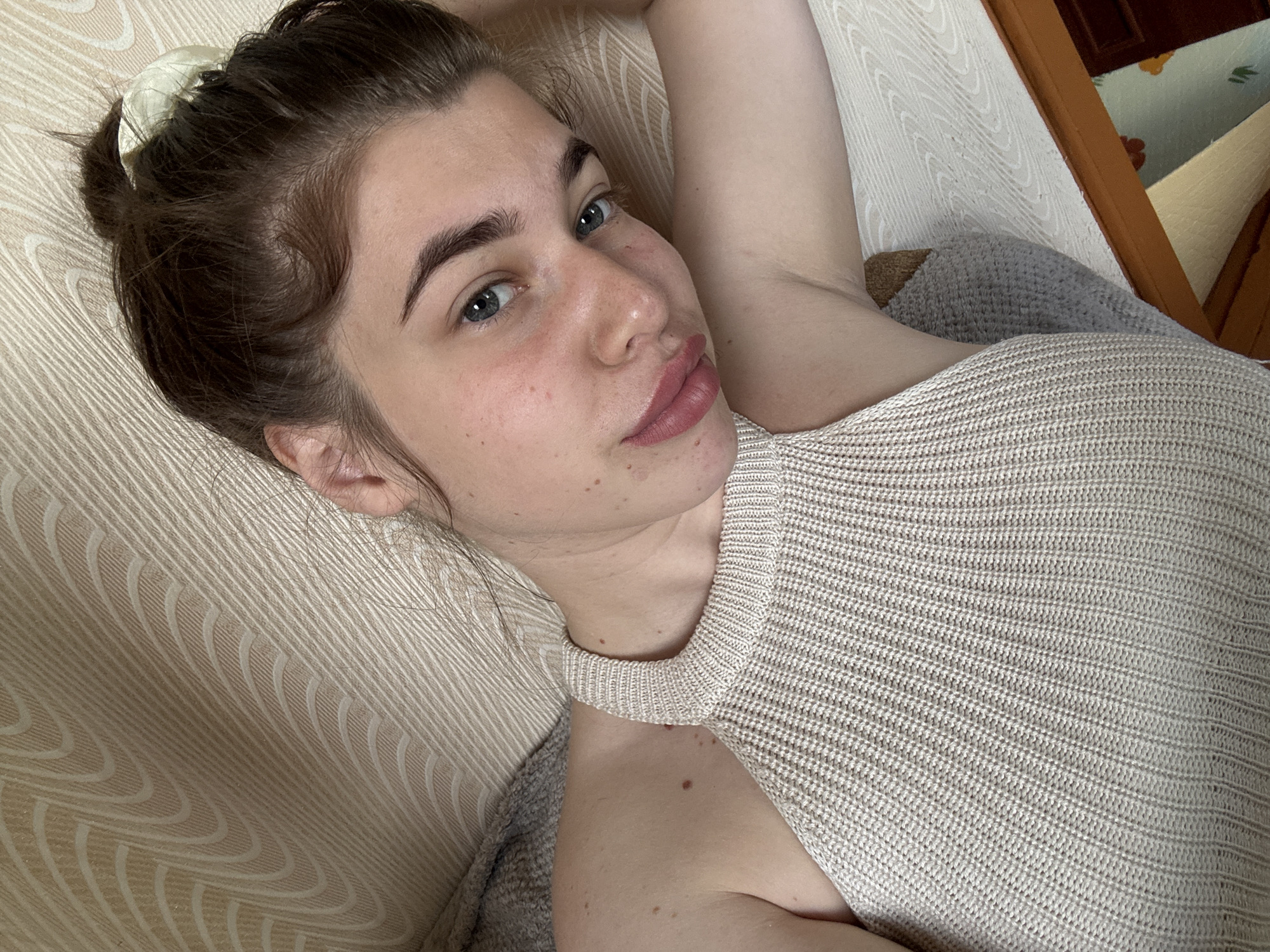 A young woman with brown hair pulled into a bun lies on a textured, beige surface, possibly a couch or bed, in what appears to be a domestic setting. She is wearing a beige, knitted halter top. Her head is tilted, and her gaze is directed slightly away from the camera. The background features a patterned wallpaper or fabric. There are no obvious indications of the specific city of Мещёры, Russia, within the image itself.