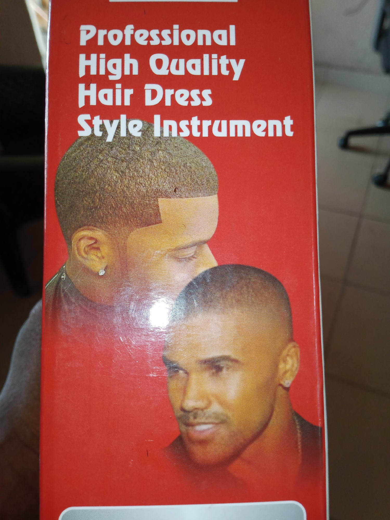 This is a close-up image of a product box with a red background. The box features white text at the top and images of two men's faces. The text reads "Professional High Quality Hair Dress Style Instrument". The faces appear to be of Black men with short haircuts. The man in the foreground has a slight smile and is looking towards the camera. The man behind him is partially visible, with his ear and side of his face showing. Both men are wearing earrings. The image is somewhat blurry and shows some signs of wear and tear, with scratches visible on the surface. The overall impression is that of a product related to hairstyling or grooming. The context provided is Jalingo, Nigeria, but this information is not directly evident from the image itself. There are no strong cues about the time of day, weather, or specific location other than the potential product context. The emotions conveyed by the men are neutral to positive, with the man in the foreground appearing to be smiling.
