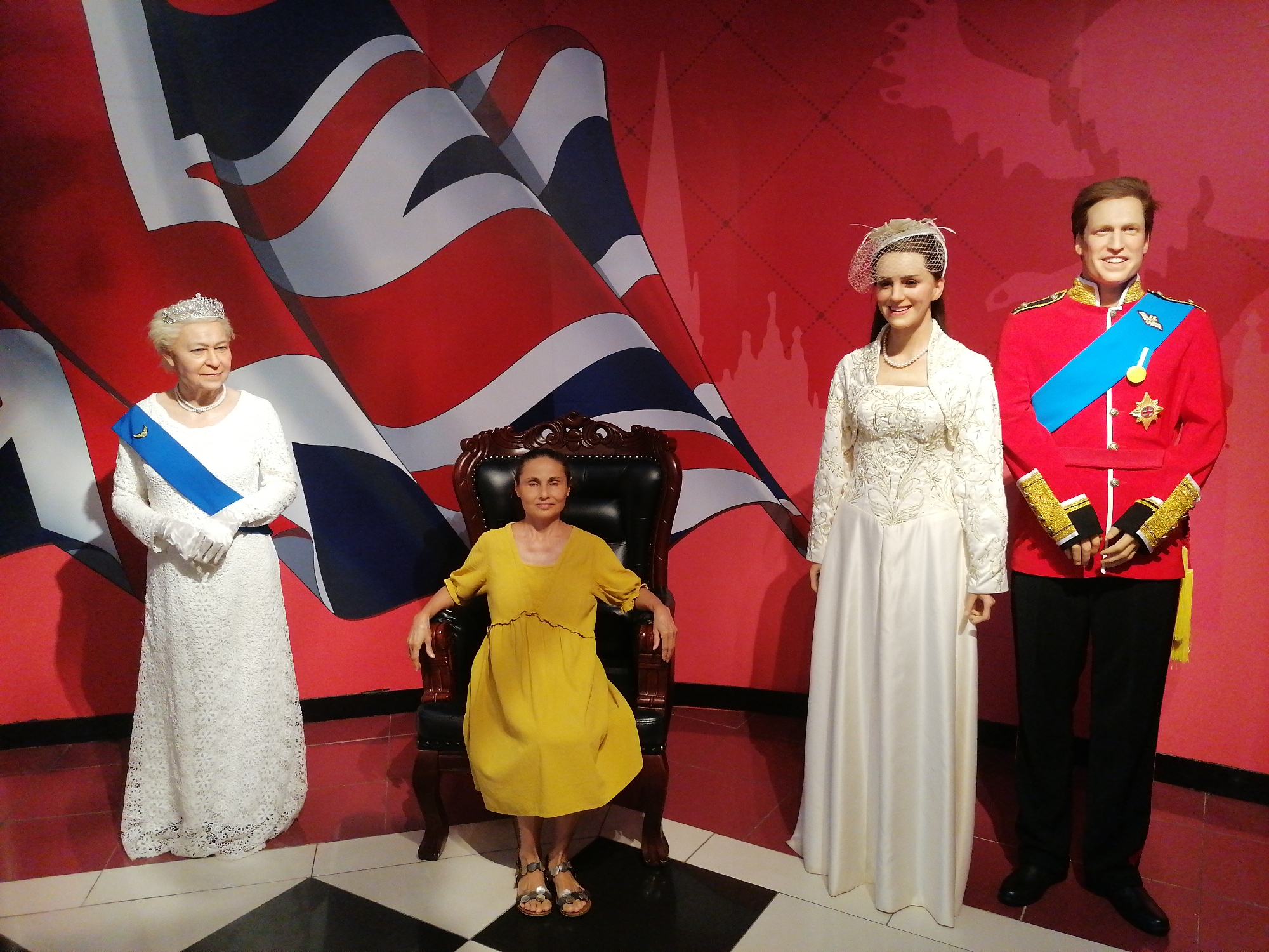 A woman in a yellow dress sits in a black leather chair, flanked by wax figures of Queen Elizabeth II and the Duke and Duchess of Cambridge. A British flag is in the background. The location is a wax museum in an unknown city.