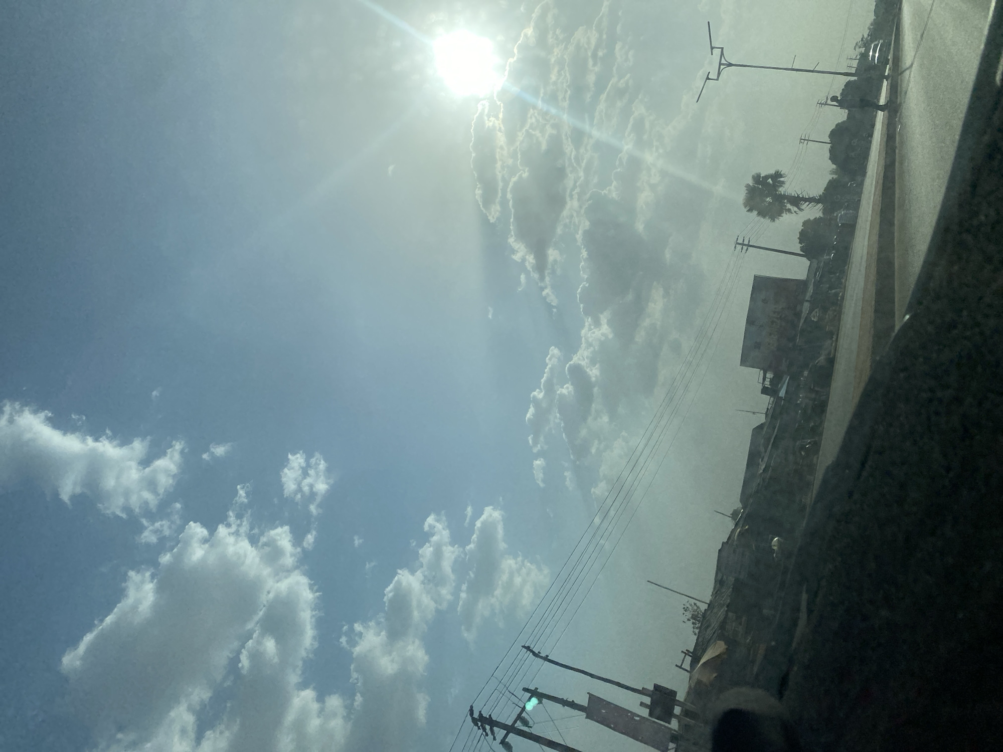The image displays an outdoor roadside scene in Sabon Tasha, Nigeria, captured from the interior of a vehicle, evidenced by a dark, angled surface in the lower left foreground. The sky is light blue with scattered white cumulus clouds, and the sun is intensely bright in the upper right quadrant, producing significant lens flare.
In the midground, a paved road with a raised median strip extends from left to right. Along the left side of the road, multiple utility poles support numerous overhead power lines; one pole exhibits a distinct green light, possibly a traffic signal, and an attached rectangular object. Adjacent to these poles are various low-rise structures, predominantly featuring corrugated metal roofs, lining the roadside. A tall palm tree is centrally located behind these structures. On the right side of the road, a modern street light pole with dual horizontal arms and solar panels is visible. An individual stands upright on the road's shoulder or sidewalk near this street light. Faint vehicular outlines are present further down the road in the distance. The scene is brightly illuminated by direct sunlight.
