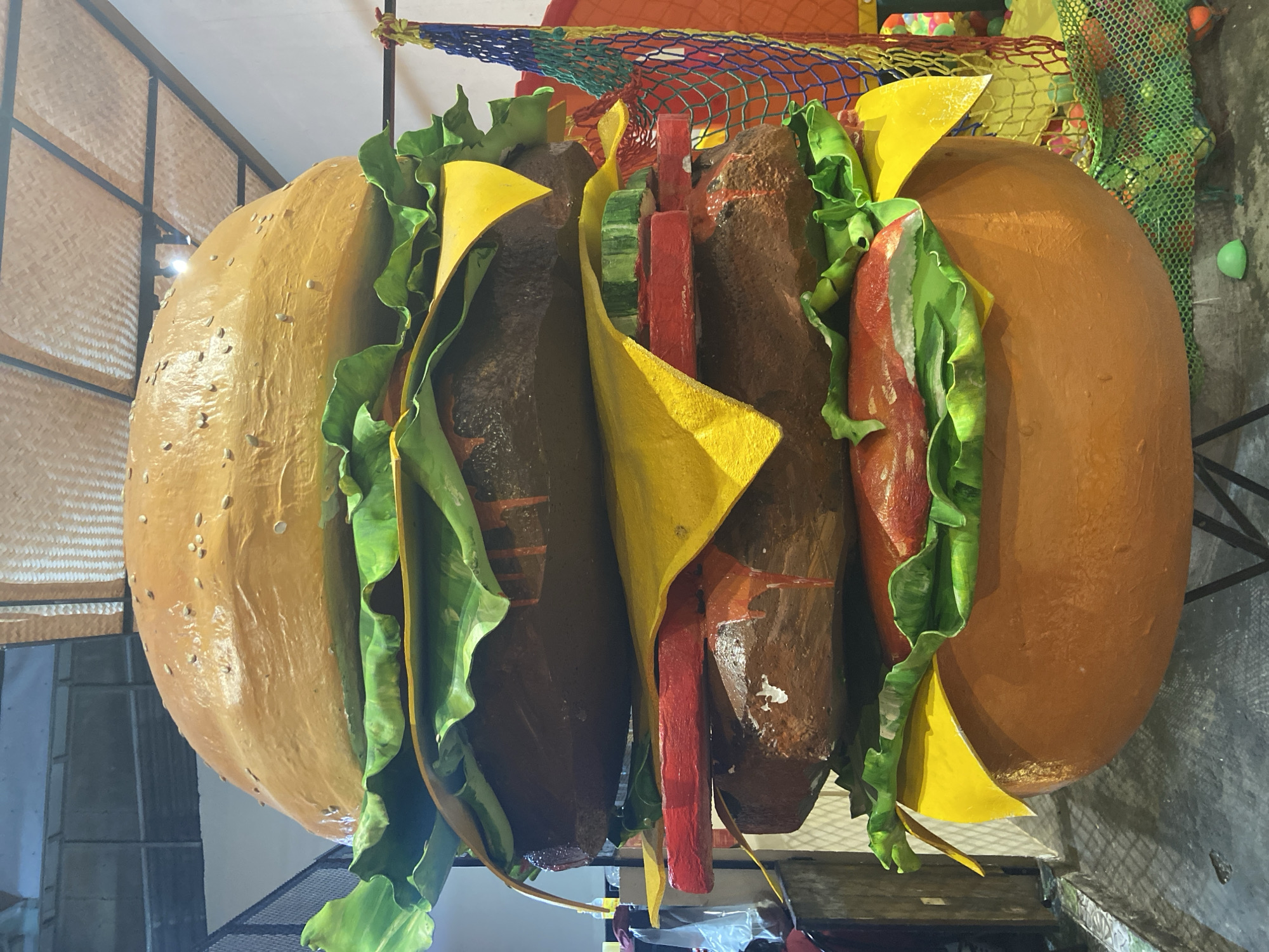 The image shows a large, vertically oriented, artificial hamburger, likely a prop or sculpture. The hamburger has two prominent buns, a deep orange-brown color, with scattered sesame seeds visible on the top bun. Between the buns, there are multiple layers representing a burger patty, lettuce, cheese, and other toppings. The patties are a dark brown and appear somewhat rough in texture. Bright green lettuce leaves with a wavy texture are visible, tucked between the patties and buns. Slices of bright yellow cheese are also strategically placed. Red and green rectangular pieces, likely representing tomatoes and pickles, are visible in the center layers. The hamburger is situated in an indoor setting, possibly a playground or entertainment area, given the presence of a colorful net with some round objects behind it and a background that suggests a wall with a woven texture. The lighting appears to be artificial, casting shadows and highlighting the glossy finish of the buns. The scene is static, with no people or animals visible. The context provided indicates the location is Nha Trang, Vietnam.