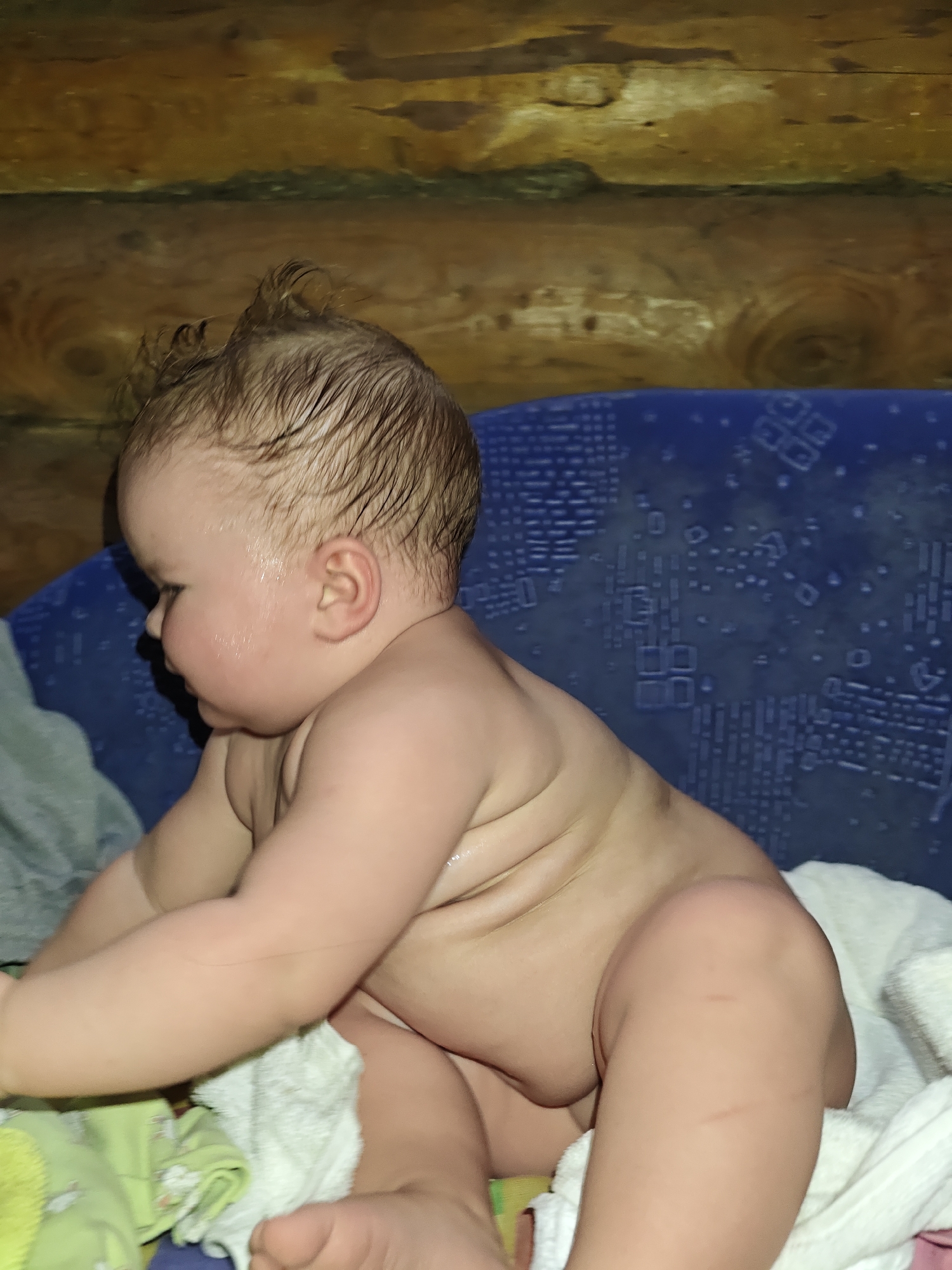 A baby is seated in an interior space with log walls. The baby has wet hair and a plump physique. A blue patterned cushion is visible behind the baby. A white towel and other green-toned textiles are present. The location is noted as Уральское, Russia.