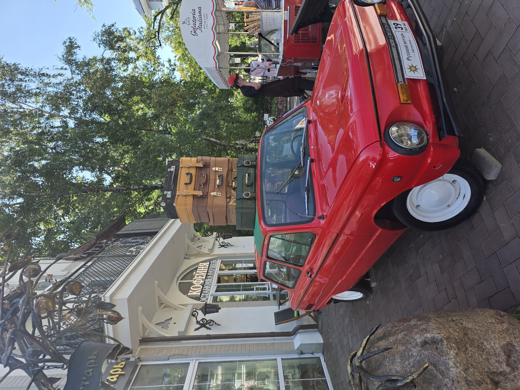 An eye-level, medium shot captures a bright red, vintage Soviet-era car parked on a brick-paved walkway in front of a building with white siding and arched windows. The car, a ZAZ 968M, has a stack of vintage suitcases, including brown leather and tan ones, secured to its roof. A green tarp is visible on top of the car, likely to protect the suitcases from the elements.
To the right of the car, an outdoor ice cream stand with a white canopy that reads "Gelateria Italiana" is set up. Several ice cream cones are displayed in a basket. A person wearing a red cap and a brown apron stands behind the counter, appearing to be engaged in a conversation on their phone. Behind the ice cream stand, a few people are visible, including one wearing a striped shirt.
The building behind the car has signs indicating it's a cafe, with Russian text that translates to "Coffee" and "Ostanovka Rayshen" (likely a cafe name or location). There's outdoor seating with chairs and a table. The scene is set against a backdrop of lush green trees and a clear blue sky. In the lower left corner, a large rock with a bronze sculpture of insect legs is partially visible, suggesting it might be an artistic installation. The overall atmosphere appears to be a sunny, pleasant day in a European-style streetscape. The license plate on the car reads "Русский янтарь 39 СВЕТЛОГОРСК RUS," confirming the location as Svetlogorsk, Russia.