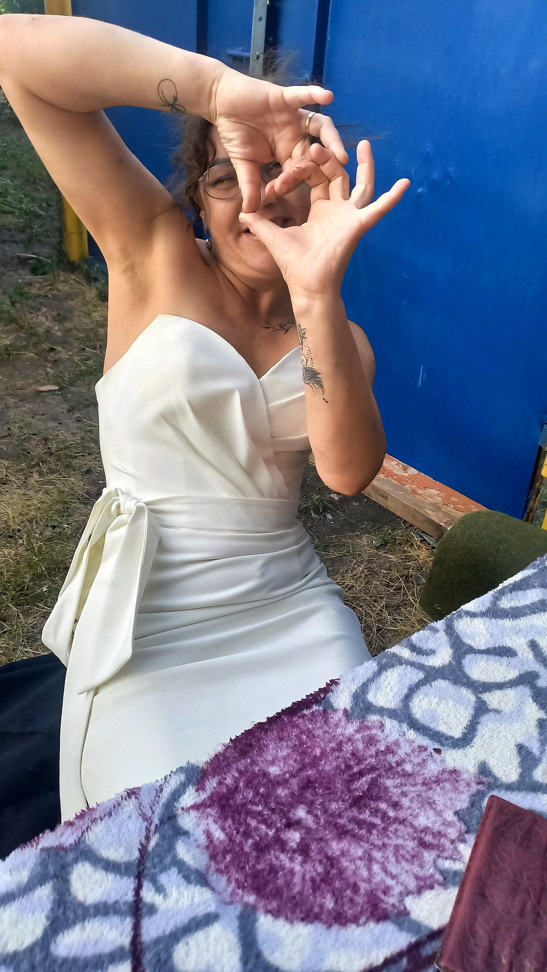 A woman wearing a white strapless dress is making a heart shape with her hands in front of her face. She is wearing glasses and has curly brown hair. She has tattoos on her left wrist and forearm. The woman is outdoors, with dry grass and dirt visible in the background. A blue structure is behind her, and a patterned blanket is in the foreground.