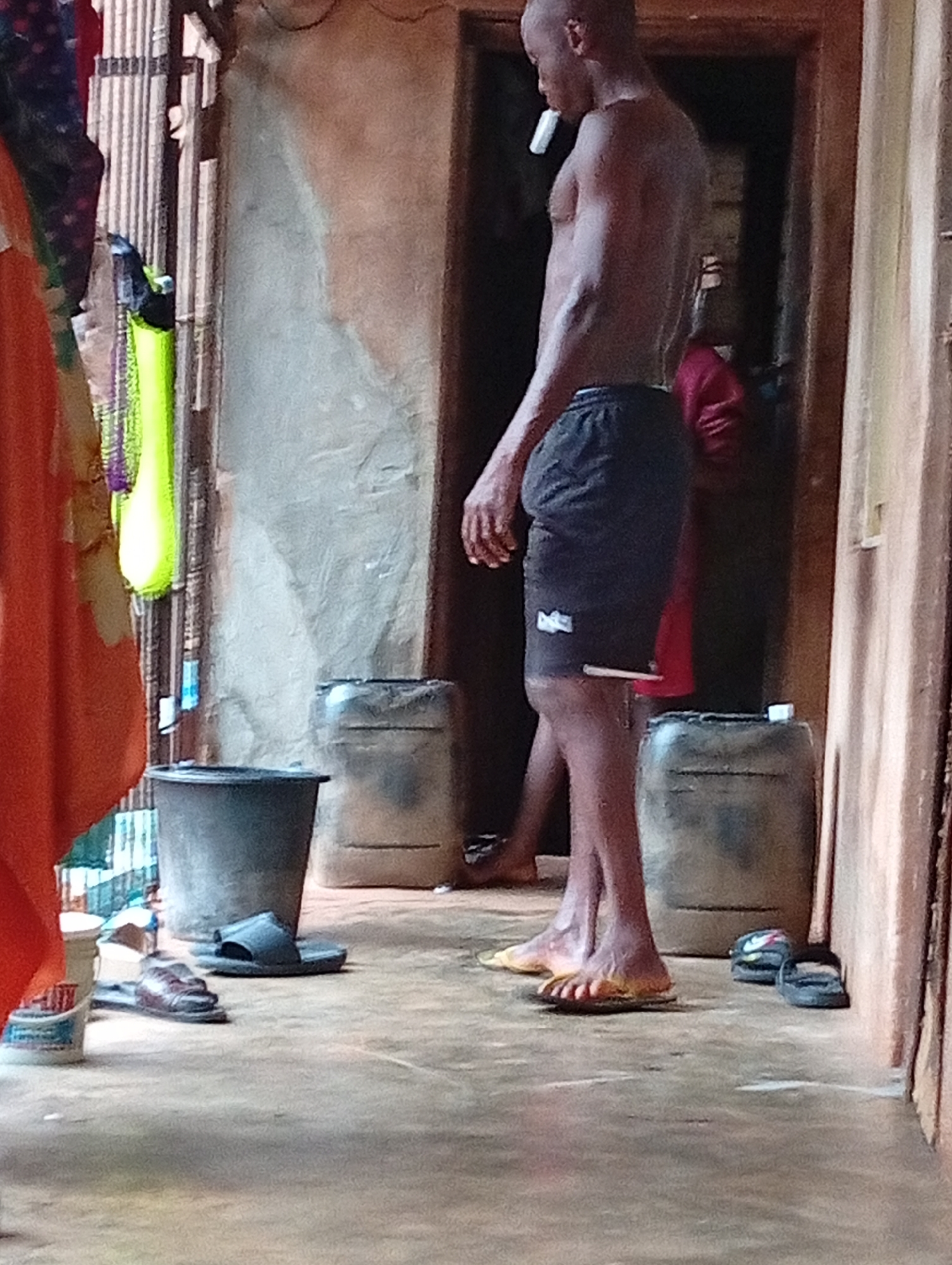 An adult African male stands shirtless in a room in Ogidi, Nigeria. He is wearing dark shorts with a white logo and has a white object in his mouth. Two other individuals are vaguely visible behind him through an open doorway. Two large water containers sit on the floor behind the man, while a dark plastic bucket is in the foreground to the left, next to a pile of sandals. Clothes hang on a rack to the left of the frame. The walls appear to be plaster.