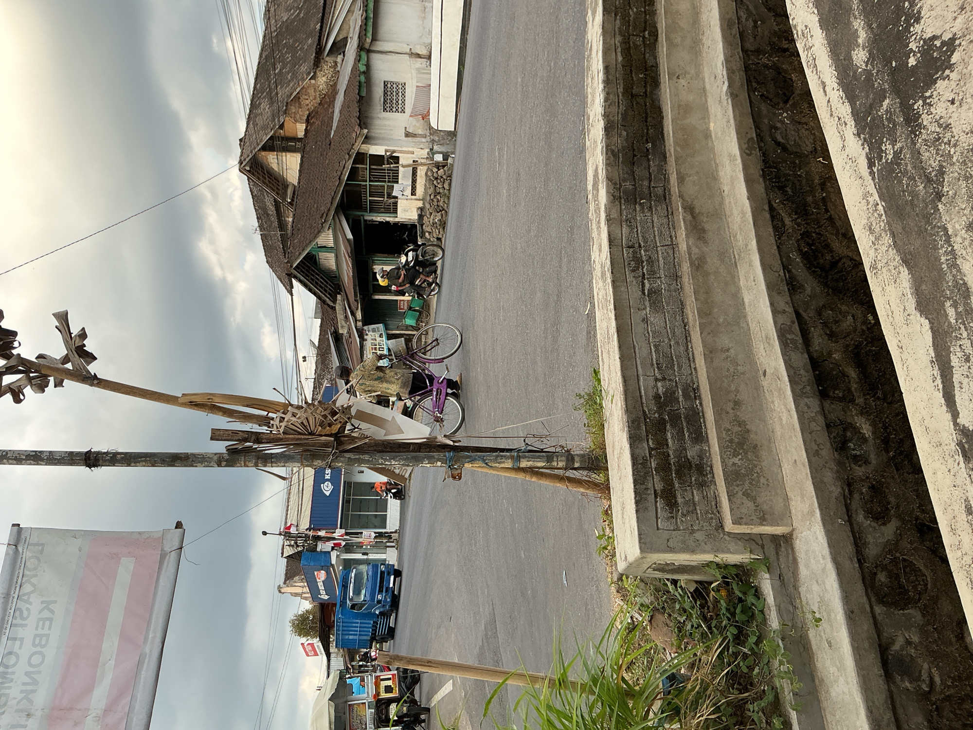 A street scene in Kebonrejo, Indonesia, shows a paved road with a drainage ditch in the foreground. A person on a bicycle with a purple frame is riding on the road. Behind the cyclist, a blue truck drives away from the viewer, and a motorcycle carrying two people approaches from the distance. Several buildings with tiled roofs line the street. A large sign is partially visible at the top left of the frame. The sky is overcast with gray clouds.