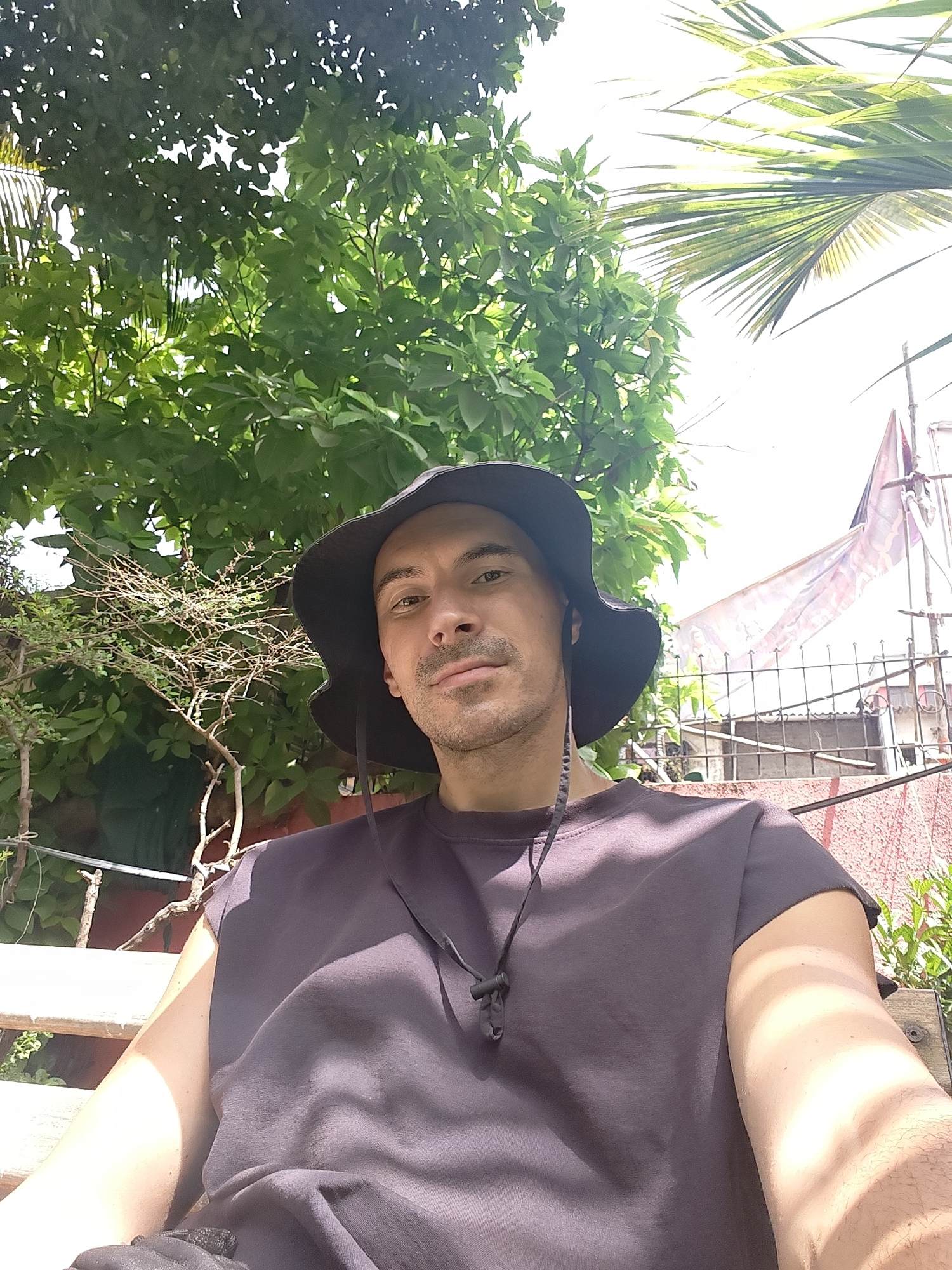 The image shows a man wearing a black hat and a dark purple sleeveless shirt, sitting outside. Behind him is a lush green tree and a palm frond, suggesting a tropical or subtropical environment. In the background, there is a fence, a building, and what appears to be a flag or cloth hanging. The exact location within an unknown city is not identifiable from the provided image.