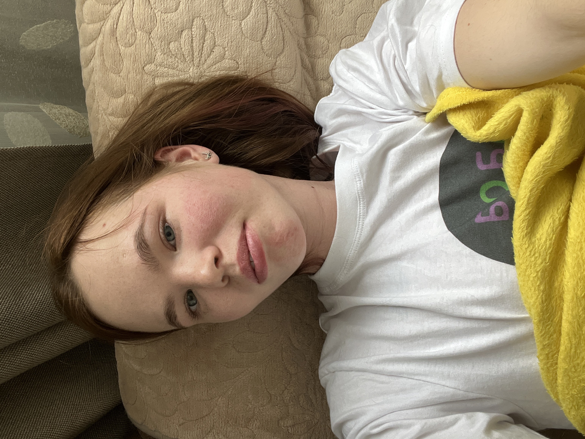 A light-skinned young woman with freckles and light brown hair is lying on a light brown patterned cushion. Her face is turned toward the camera. She is wearing a white t-shirt with a graphic design of a dark gray circle with some pink and green design on it. A yellow cloth covers part of the shirt. The background is nondescript. The location cannot be determined to be Chaikovsky, Russia, based on the visual information provided.