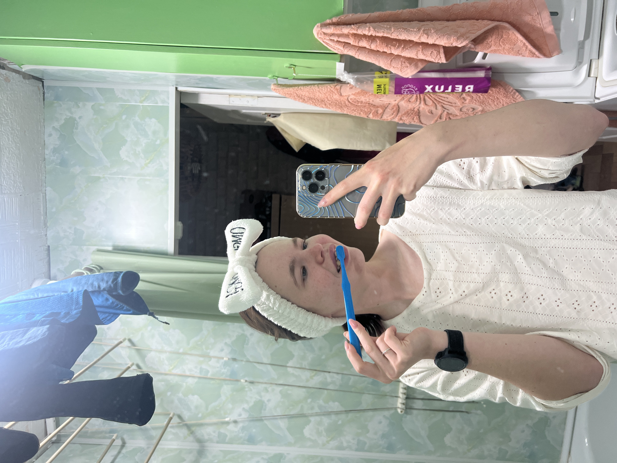 A young woman in a white lace shirt and white bunny-ear headband is in a bathroom, using a blue toothbrush to brush her teeth while holding a smartphone up to capture her reflection in a mirror. The mirror reflects the bathroom scene, including a green cabinet above the mirror and a peach-colored towel hanging on the cabinet door. A package of 'BELUX' tissues is visible on the cabinet. To the left of the woman, a clothes drying rack holds various items of clothing, including a blue jacket and black trousers. The bathroom walls are covered with a light green patterned tile. Based on the location provided, this scene likely takes place in a bathroom in Chaikovsky, Russia.