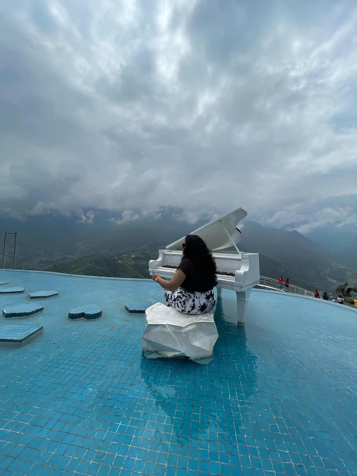 A woman is sitting on a rock in the foreground, facing away from the camera, with her hands resting on the keys of a white grand piano. The piano is positioned at the edge of a bright blue tiled infinity pool. In the background, rolling green hills and mountains are shrouded in a dramatic, cloudy sky. Several other people are visible on a walkway in the mid-ground, and a few unique, heart-shaped blue platforms are scattered in the pool. The scene appears to be at a scenic overlook or resort in Sa Pa Ward, Vietnam, known for its mountainous terrain.