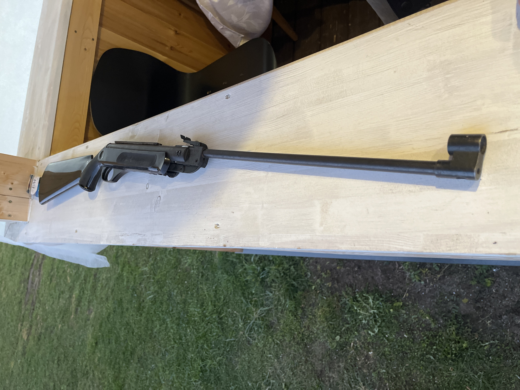 The image shows a black air rifle lying on a white wooden surface. The rifle is complete, with its stock resting near the edge of the surface. The surface appears to be part of a wooden structure. In the background, there are indistinct objects, including what appears to be a black musical instrument case and a white plastic bag. The background also includes wooden planks. The bottom of the image shows a grassy area with some dirt. The location is Гаревая, Russia.