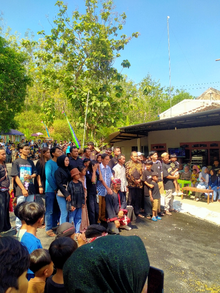 The image depicts an outdoor gathering in Tawangsari, Indonesia, under a clear blue sky on a sunny day. A large group of approximately 20-25 adults and several children stands in the midground on a dark, possibly paved, surface. This central group includes individuals of varying ages, dressed in a mix of casual attire such as t-shirts, jeans, and hoodies, with some wearing more traditional or semi-formal clothing, like a man in a brown and gold batik shirt. One man in the foreground of this group is kneeling, wearing a dark top, dark trousers, and a distinctive red and white patterned head covering. Another man on the far right of the standing group holds a young child.
In the background, lush green foliage and tall trees are visible. To the right, a single-story building with a light-colored exterior and a dark roof overhang is present. A tall, slender antenna or pole rises vertically above this building. Inside the building, electronic equipment including what appears to be a television and a stereo system are visible. Further to the right, a smaller group of people, primarily women and children, are seated on green plastic chairs and a wooden bench.
In the immediate foreground, the backs of several children's heads, including one wearing a green hijab, partially obscure the view. The ground directly in front of the central standing group is dark with scattered yellow-orange granular debris, possibly corn kernels or flower petals. Decorative vertical elements in green and blue are visible behind the standing crowd on the left.