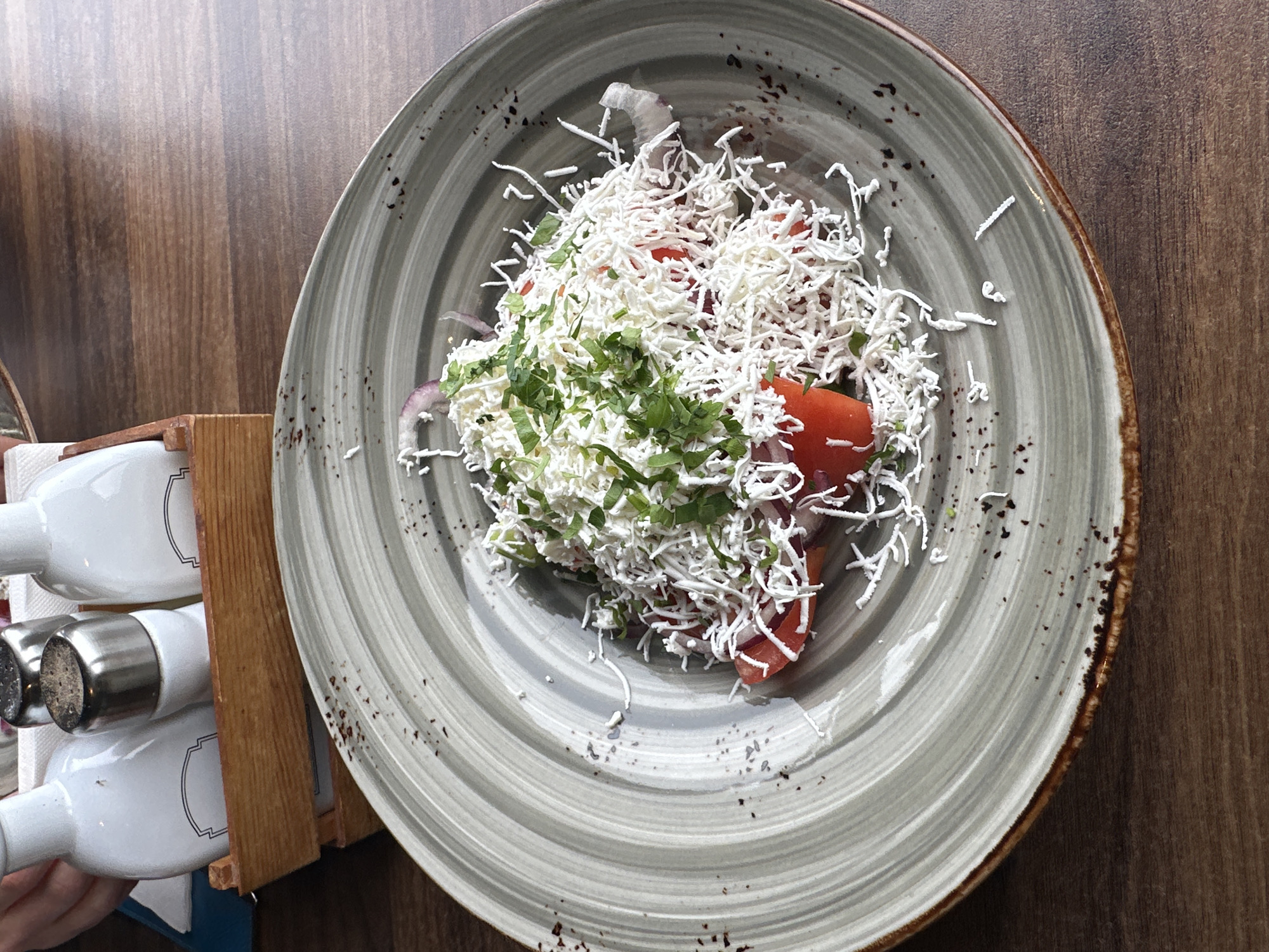 Here's a detailed description of the image:
**Scene:** A close-up, overhead shot of a Shopska salad served on a gray and white patterned plate with a brown-rimmed edge. The plate sits on a wooden surface.
**Subjects/Objects:**
* **Shopska salad:** The salad is composed of chopped tomatoes, red onion, green peppers, grated sirene (a Bulgarian cheese), and parsley. The grated sirene covers most of the vegetables.
* **Plate:** A round plate with concentric circles in shades of gray and white. It has a slightly raised rim with brown detailing.
* **Wooden Table:** A brown wooden surface on which the plate is placed.
* **Condiment Set:** A wooden holder containing white bottles, most likely for oil and vinegar, with metal tops and shakers. The holder also appears to have some tissues.
**Environment:** The setting appears to be a restaurant or cafe in Vidin, Bulgaria. This is inferred from the presence of a typical Bulgarian dish (Shopska salad) and the overall presentation suggests a dining establishment.