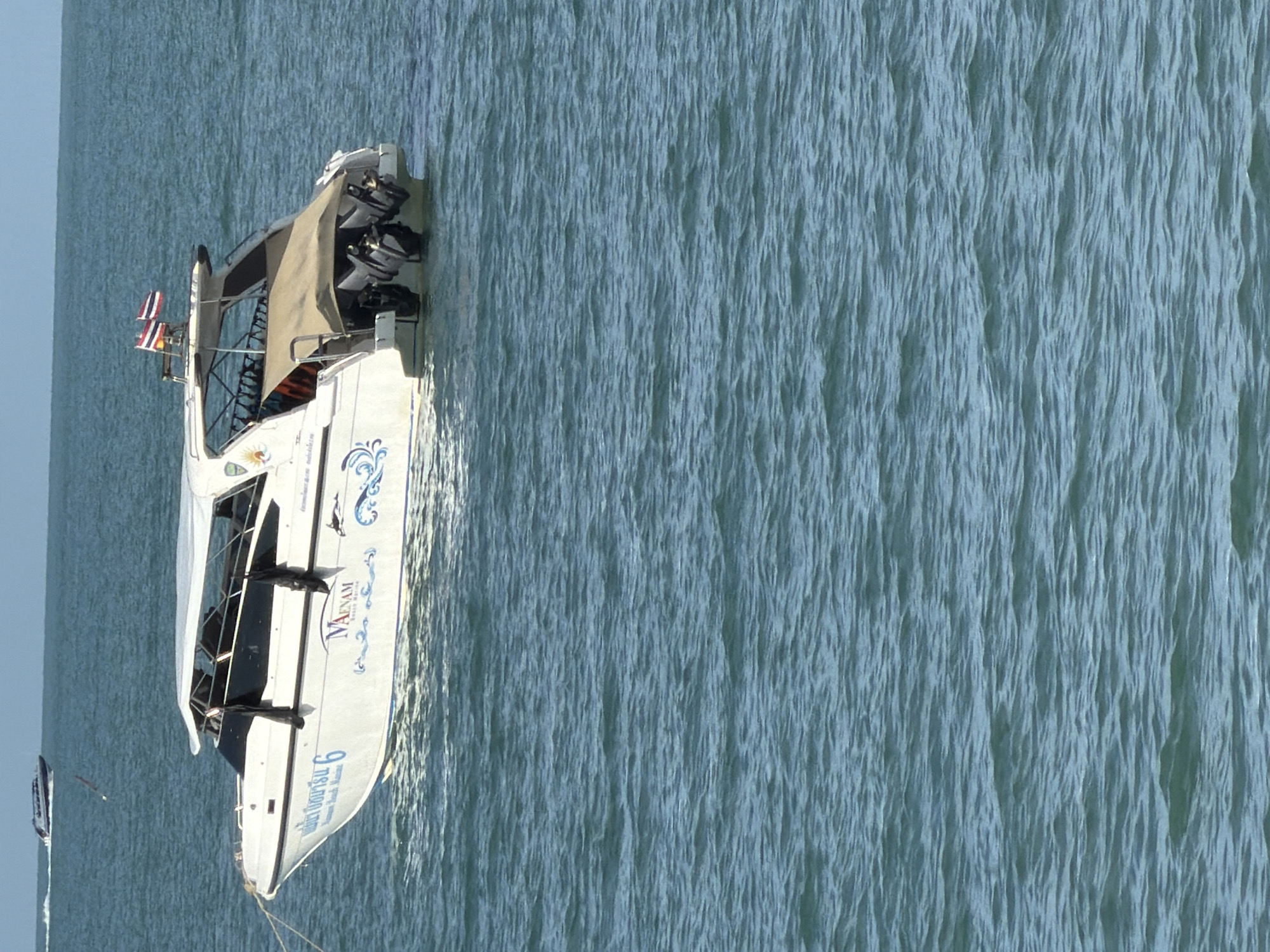 A white speed boat with a blue stripe along its hull is anchored in calm, blue ocean water. The boat has a covered passenger area with black windows and a Bimini top. Two Thai flags are visible at the stern of the boat. There are three outboard motors mounted on the back of the boat. Text in Thai and English is visible on the side of the boat, including "MAENAM" and the number "6". The scene is likely in or near Baan Tai, Thailand, given the boat's name, which refers to Maenam Beach on Koh Samui, a popular tourist destination near Baan Tai. The sky is clear and a light blue. Another boat, a darker color, is visible in the distance on the horizon.