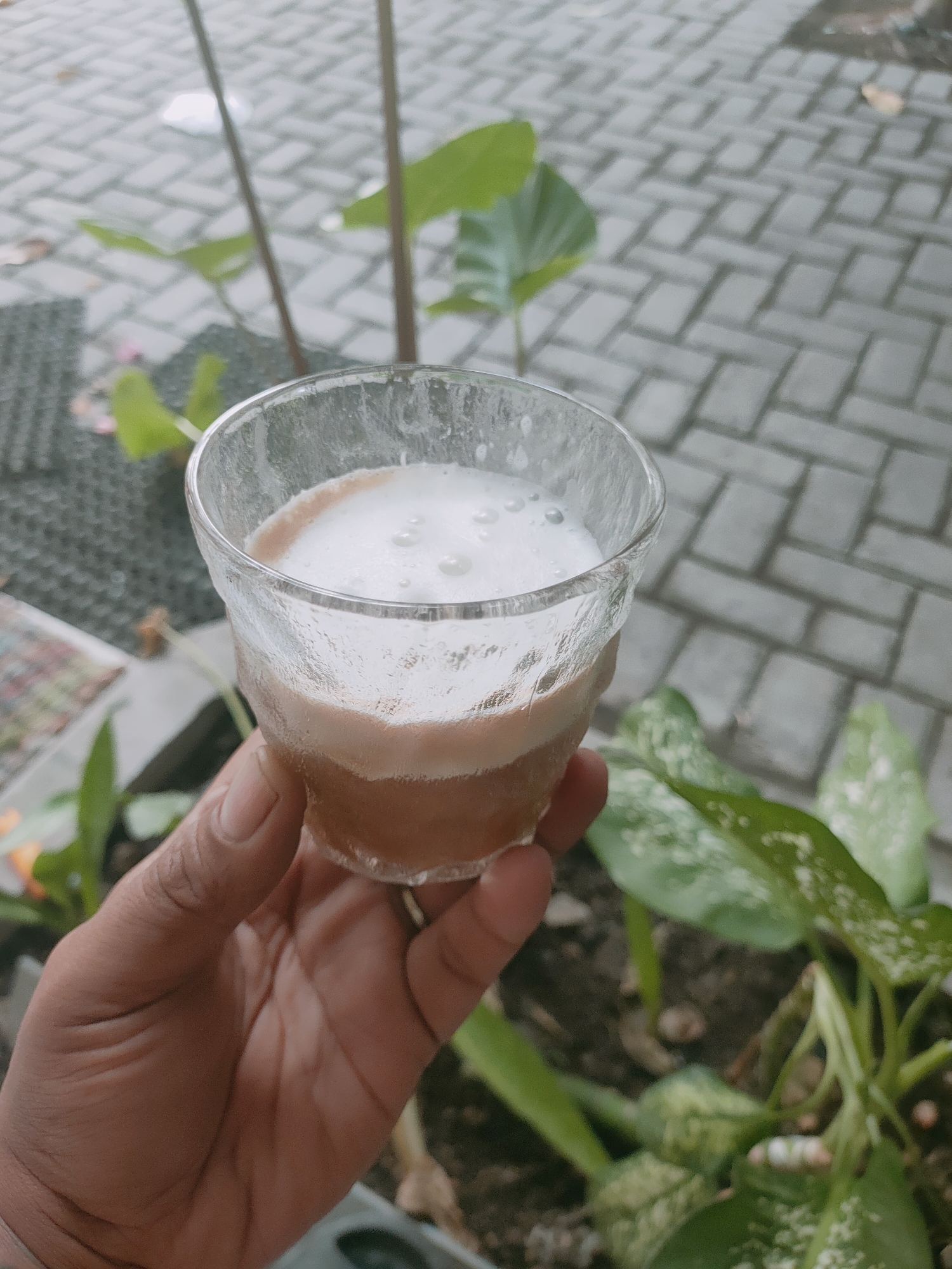 A hand holds a clear glass containing a frothy beverage. The glass is cylindrical with a textured surface. The beverage is layered, with a lighter, foamy top and a darker liquid below. The background is composed of a grey, patterned pavement. Green foliage and a few dry leaves are visible in the midground and background, suggesting an outdoor setting. The scene appears to be in the Sooko area of Indonesia.