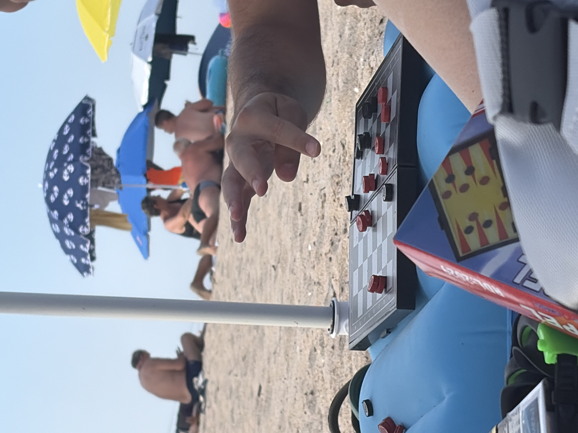 An adult's hand hovers over a checkerboard set up on an inflatable mat on a sandy beach. The hand is positioned to make a move, with fingers extended towards the board. The checkerboard is partially visible, with red and black checkers in place. In the background, several other beachgoers are seen sitting or lying on the sand, some under beach umbrellas. The sky is clear and blue. The overall scene depicts a leisurely day at the beach.
