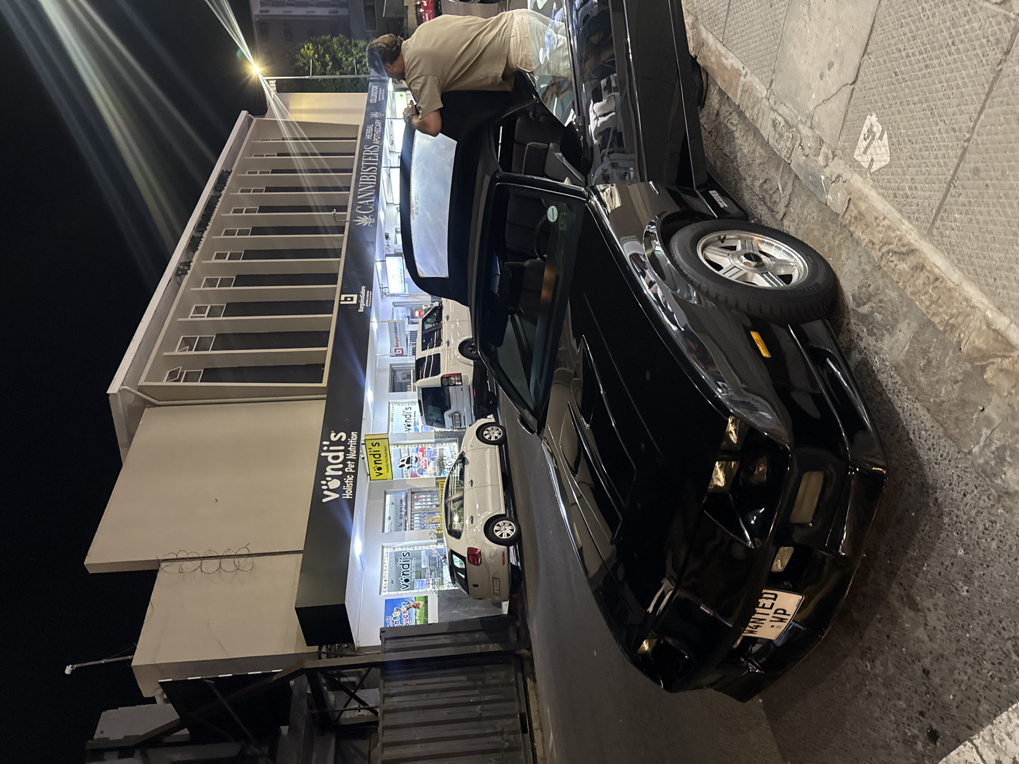 The image shows a black vehicle parked on a street at night in front of a building with several businesses. A person is leaning into the open driver's side of the car. The building hosts a "CANNIBISTERS" herbal apothecary, "vöndi's" holistic pet nutrition, and "BargainsGalore". Cars are parked behind the black vehicle. The license plate reads "WANTED WP", indicating that this is Cape Town, South Africa. There is a sidewalk next to the vehicle. Overhead lights illuminate the scene.