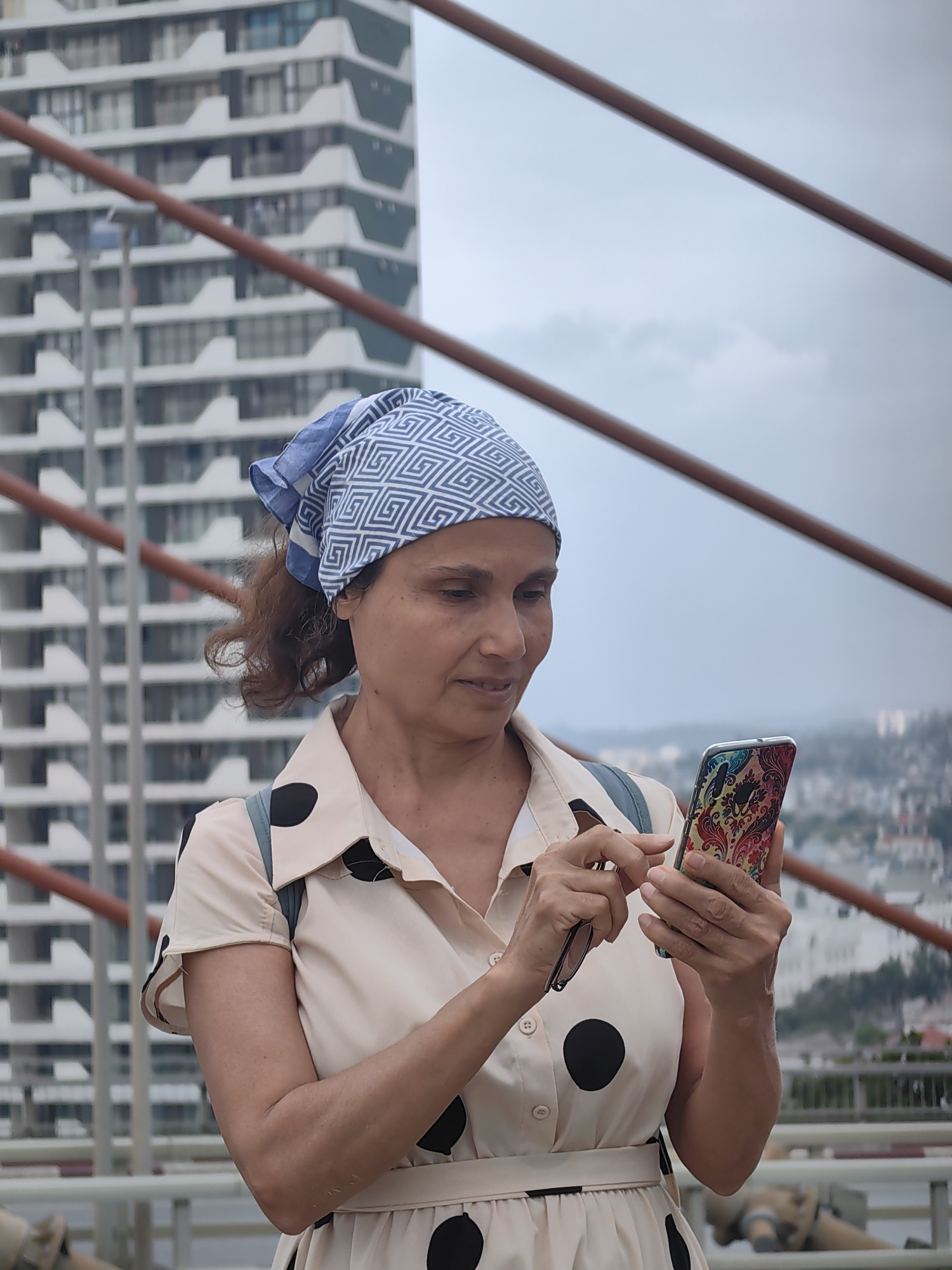 A woman stands on a bridge, holding a smartphone in her right hand and sunglasses in her left. She is fair-skinned with brown hair partially covered by a blue and white patterned headscarf. She wears a light beige, knee-length collared dress with black polka dots and a matching belt. Behind her, a tall, white building with geometric, stepped architecture is visible. The bridge has orange railings. In the background, the cityscape of Bai Chay Ward, Vietnam, is partially obscured by a hazy sky.