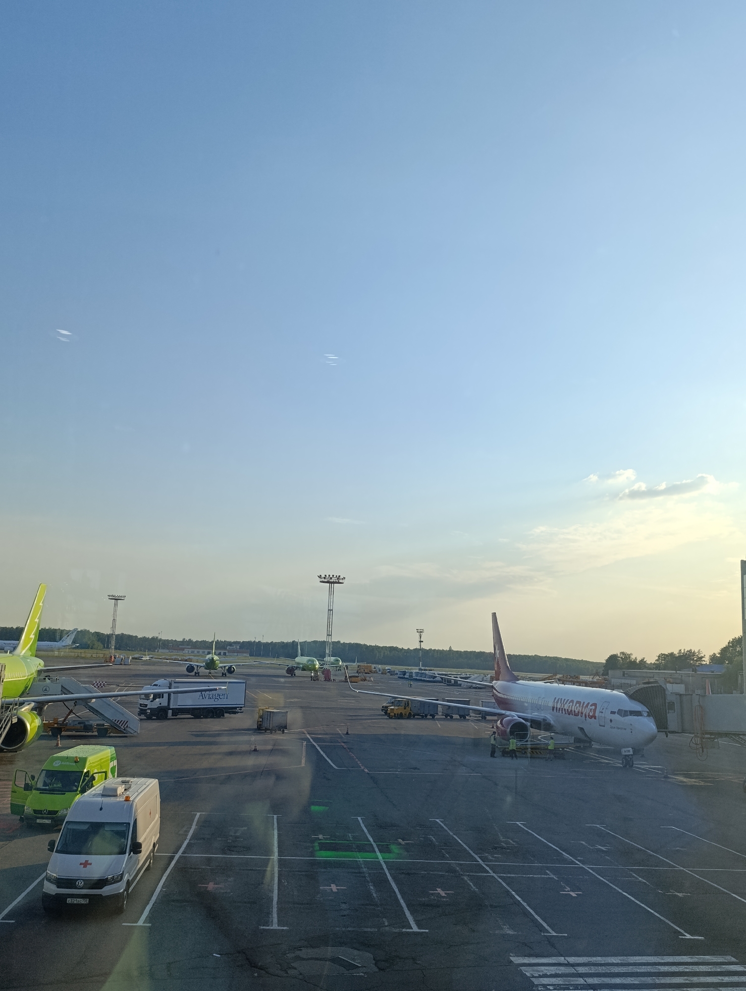 The image depicts an aerial view of an airport tarmac in Domodedovo, Russia. The foreground is dominated by a white ambulance, identifiable by its red cross emblem and license plate "C921XC 774". Adjacent to it is a green cargo van with the "Aviagen" logo. Further back, a large white truck with the "Aviagen" logo is parked. Several airplanes are visible on the tarmac, including a prominent red and white passenger jet with Cyrillic lettering on its fuselage, identified as "ИКАР" (IKAR). Another green airplane is parked on the left, and other aircraft are scattered across the airfield in the background. The tarmac is marked with white lines, indicating parking spaces and taxiways. The sky is a clear blue with a few wispy clouds, and the overall lighting suggests it is daytime, likely late afternoon due to the warm tones in the sky. The environment appears to be an active airport, with ground crew members attending to the aircraft.