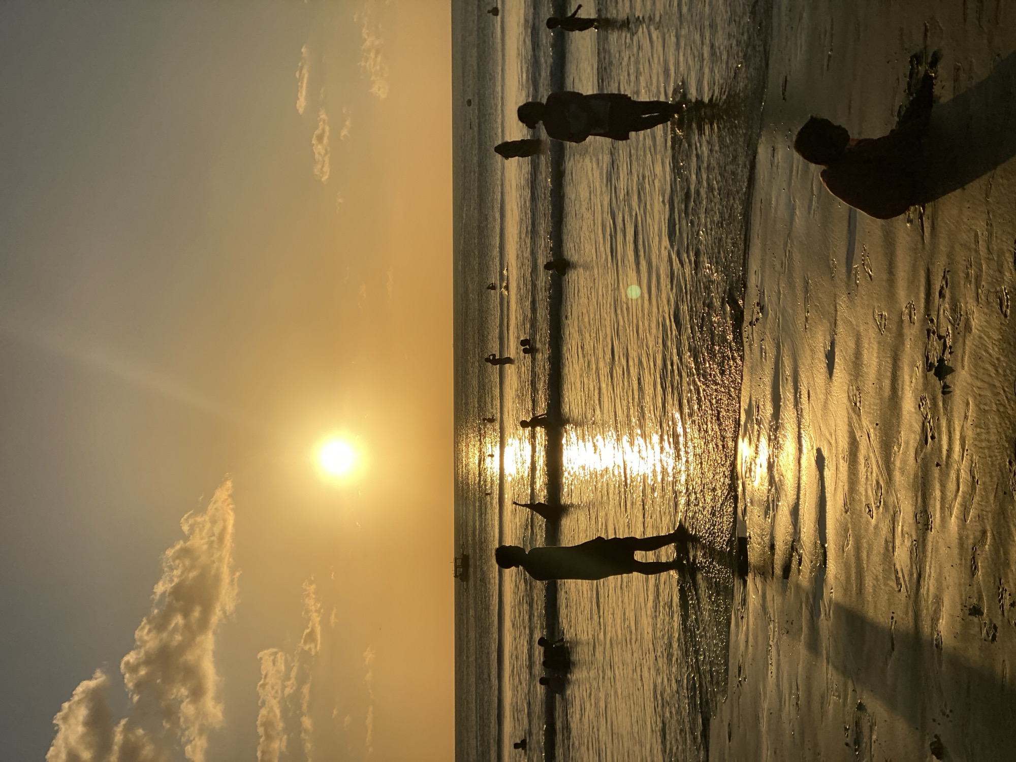 This is a photograph taken at sunset on Kuta Beach, Kuta Selatan, Indonesia. The sun, a bright orb, is low on the horizon, casting a warm, golden glow across the sky and reflecting on the water. The sky is a gradient of orange and yellow, with a few scattered clouds tinged with pink.
In the foreground, the wet sand of the beach shows ripples and footprints. A child is sitting on the sand in silhouette, appearing to play or dig. Further out, the shallow water of the ocean laps at the shore. Several people are visible in the water, silhouetted against the bright sunset. One man stands in the water, looking towards the sea. A woman and child are also in the water, close to the shore. Other figures can be seen further out in the ocean, some appearing to be swimming or playing.
The ocean stretches out to the horizon, with gentle waves breaking. A small boat is visible in the distance, on the calmer waters of the sea. The overall impression is of a peaceful and serene beach scene at the end of the day.
