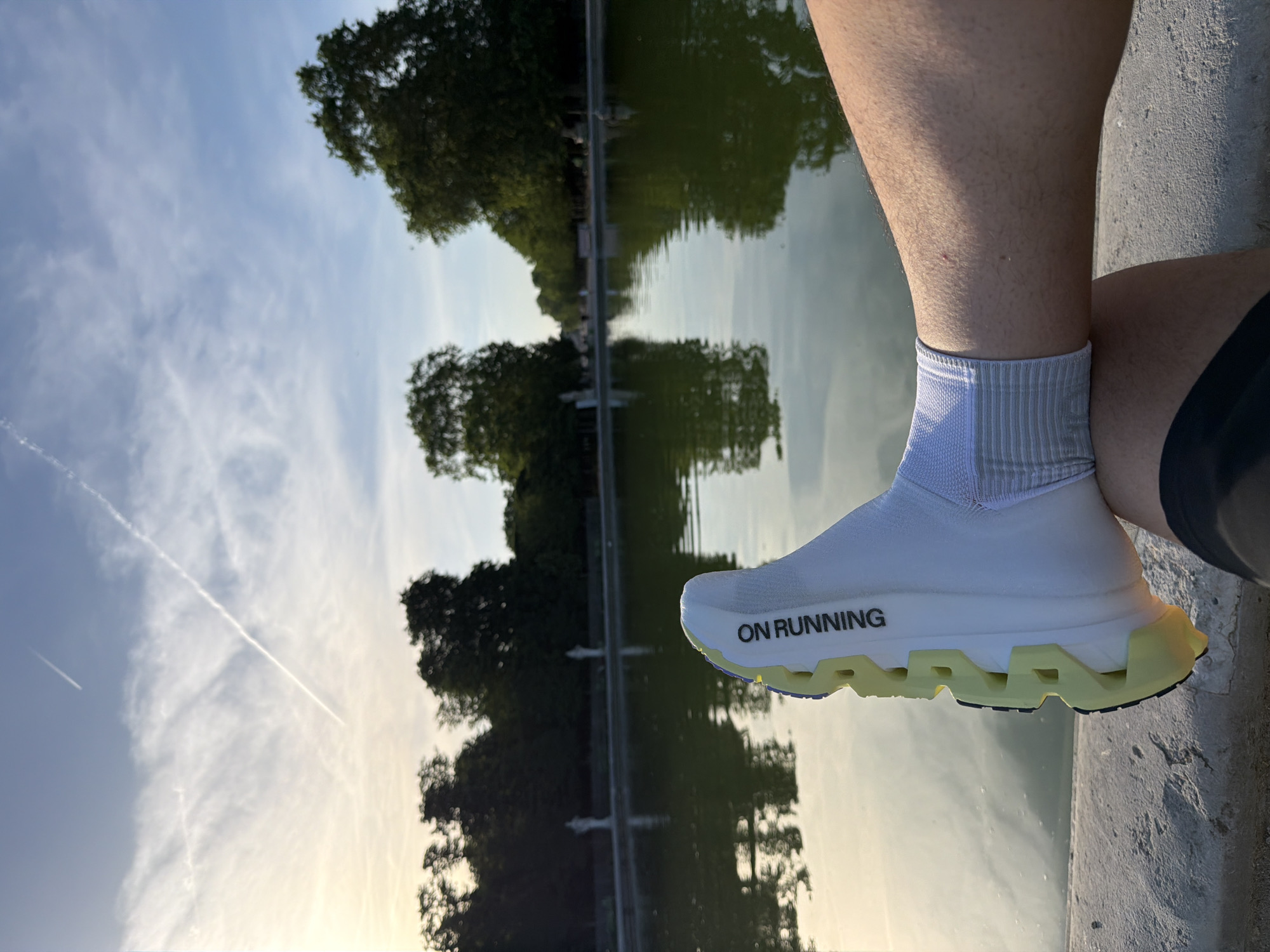 A low-angle, eye-level shot depicts a person's right foot and lower leg positioned on a concrete ledge, with the background revealing a reflective body of water mirroring trees and the sky. The shoe is a white "On Running" brand sneaker with a distinctive yellow sole. The person is wearing a white sock with gray stripes around the ankle. The background features a pond or canal reflecting the surrounding trees and a cloudy sky, suggesting a location in a park. Given the architectural style and ambiance, the scene is likely set in Paris, France, specifically in one of the city's parks featuring canals or ponds, such as the Jardin des Tuileries or Jardin du Luxembourg.