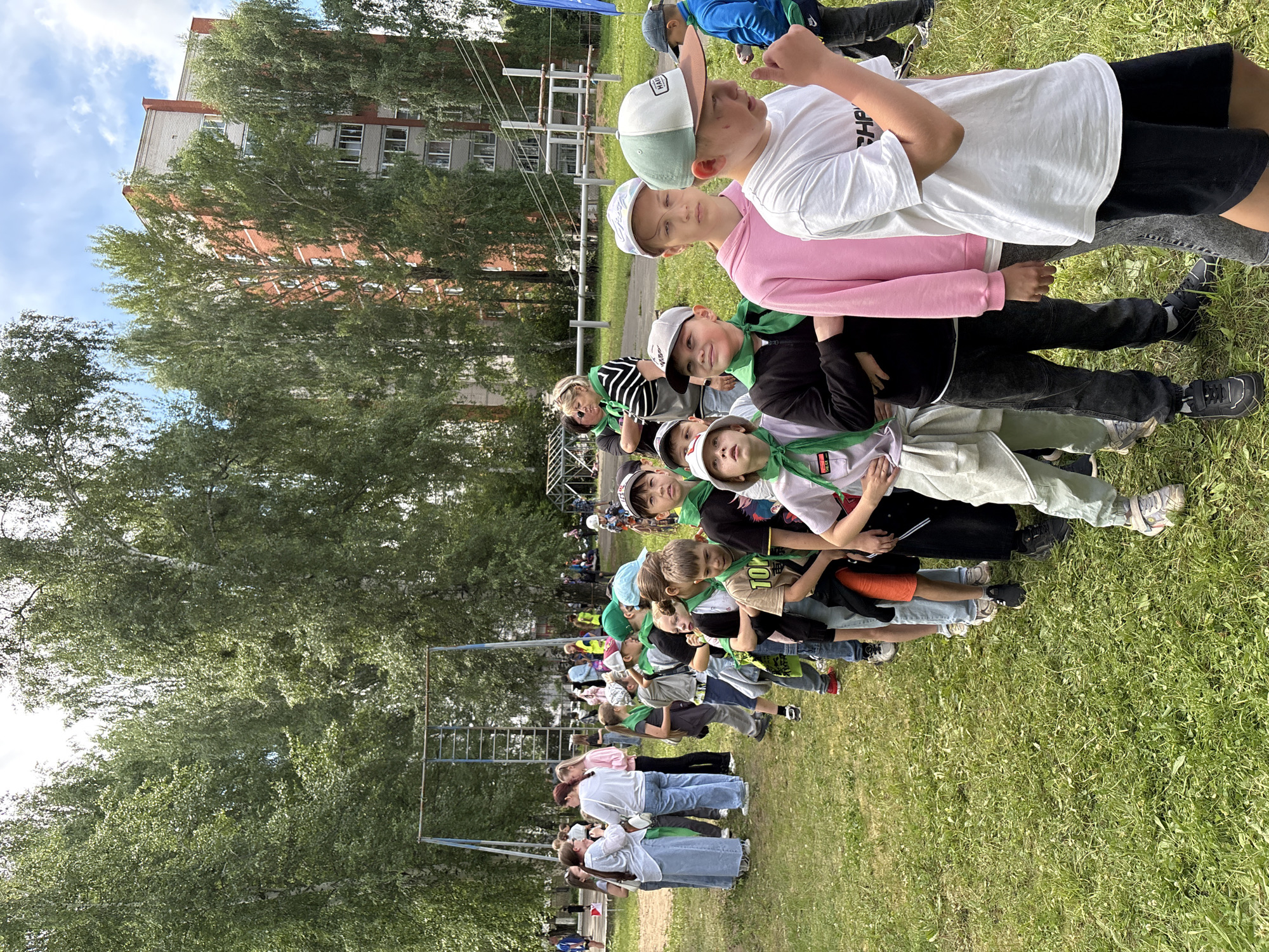 The image depicts a group of children and adults in a line on a grassy field, with trees and a multi-story brick building in the background. The children are mostly wearing casual clothing and some have green bandanas around their necks, suggesting an organized activity. There appears to be a playground structure in the mid-ground. The building in the background is a residential high-rise, typical of Soviet-era architecture. The overall impression is a community event or outdoor activity taking place on a bright, cloudy day. This scene is consistent with the urban landscape of Chaikovsky, Russia, which has many residential areas with green spaces and Soviet-style architecture.