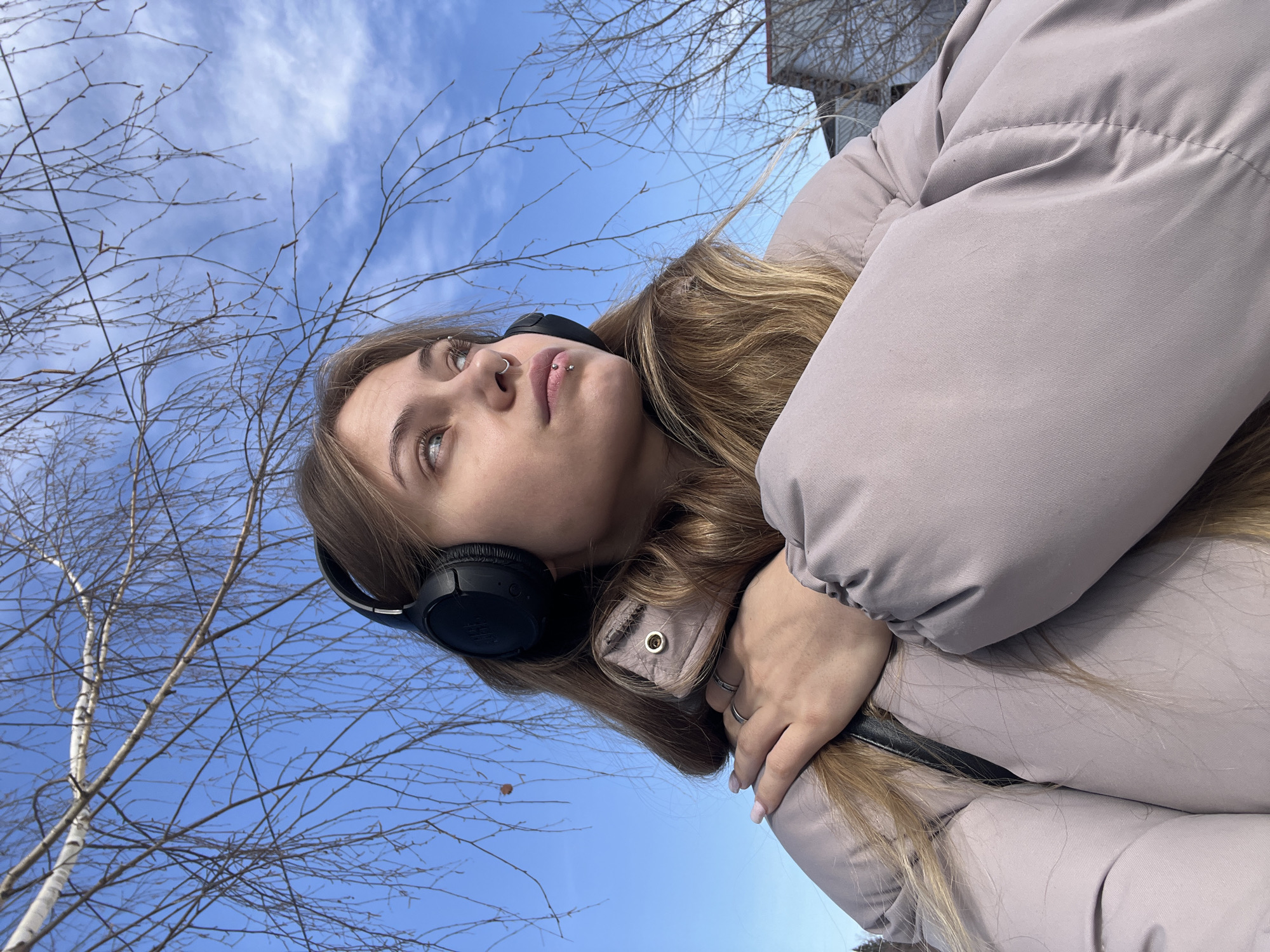 A low-angle outdoor portrait features a young adult female under a clear blue sky. She is light-skinned with long, light brown hair, wearing black JBL over-ear headphones and a light gray-beige puffer jacket. Her left hand, adorned with a silver ring on the ring finger and light pink manicured nails, rests on her right shoulder. She presents a silver hoop septum piercing, a stud above her left eyebrow, and a silver barbell piercing on her lower lip. Her gaze is directed upwards and to her left (viewer's right). The background primarily consists of bare tree branches, consistent with a cold season, and a distant building partially visible through the leafless trees. The precise location within the city of Фоки, Russia, is not ascertainable from the visual information provided.