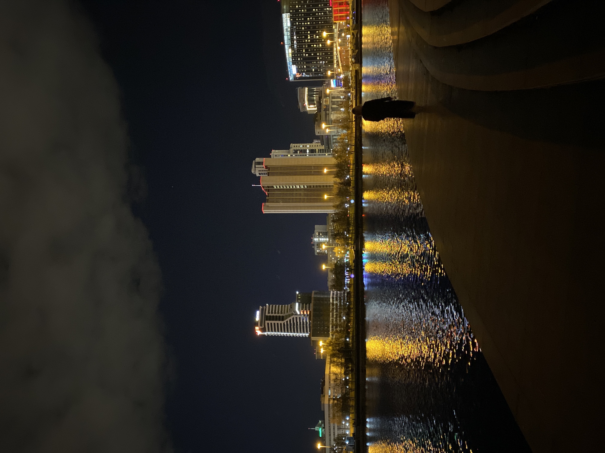 At night, the cityscape of Yekaterinburg, Russia, reflects in a body of water. Tall buildings are illuminated, and their lights cast shimmering reflections. A person stands on a structure near the water's edge, gazing at the cityscape. Cloud cover is visible in the night sky.