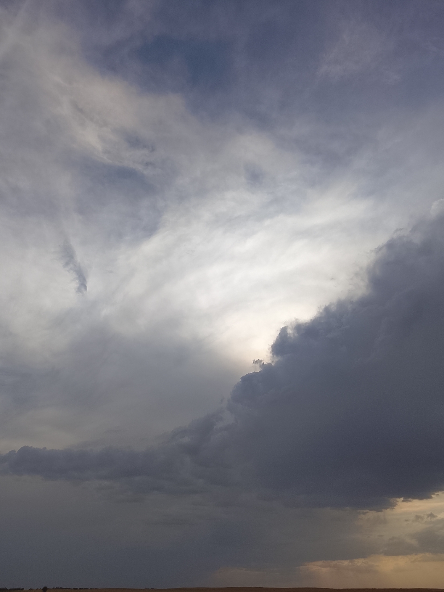The image depicts a dramatic sky with heavy, dark clouds dominating the lower right portion, partially obscuring a bright, diffused light source that appears to be the sun. Wispy, lighter clouds streak across the upper and left portions of the sky, with hints of blue visible in the far upper left. The lower third of the image shows a flat, arid landscape, possibly fields or plains, under a muted, overcast sky. The overall impression is one of impending weather, possibly a storm, with a stark contrast between the dark clouds and the bright sky. The scene is located in Stepnoe, Kyrgyzstan, though no specific landmarks or human activity are visible in this general landscape shot.
