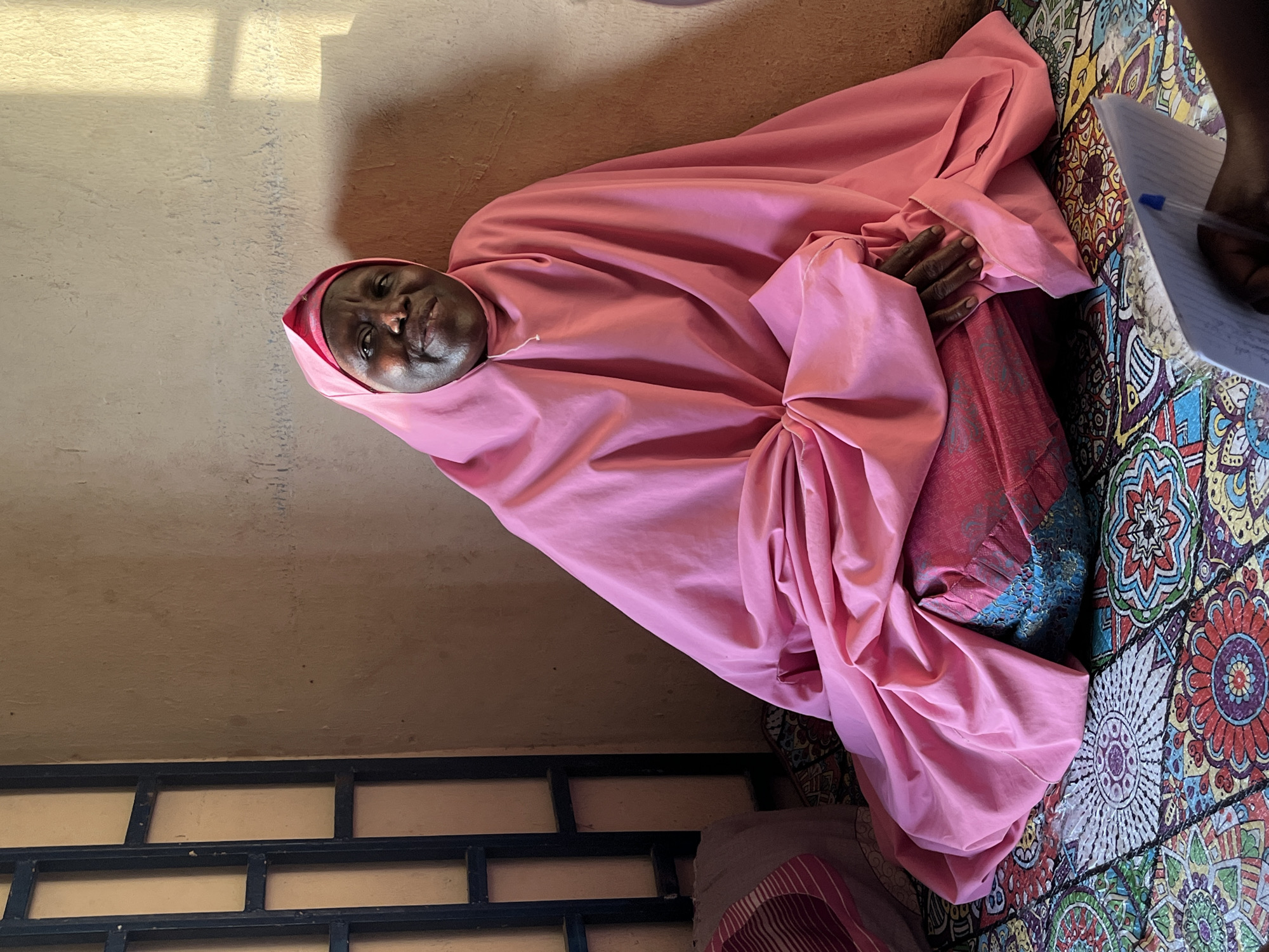 An elderly Black woman is seated indoors on a brightly colored, patterned rug. She is draped in a pink garment covering her head and body. The woman's hands are clasped in front of her. To the right of the frame, an arm is visible holding a pen and notepad. The background consists of a wall painted in light colors and a dark metal grate in the lower-left corner. This photograph was taken in Jos, Nigeria.
