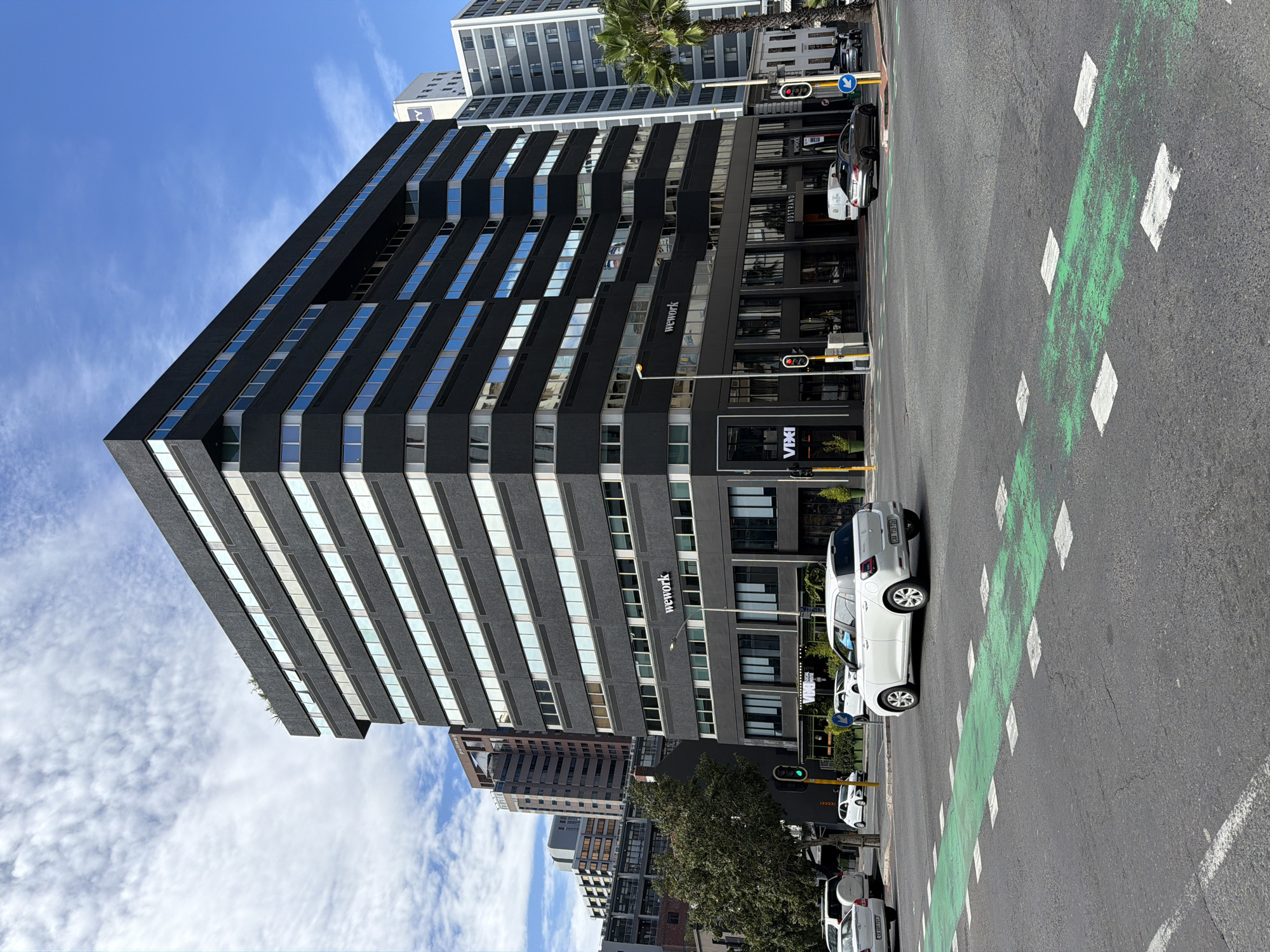 A medium, long-shot, daytime photograph shows a street scene in Cape Town, South Africa. A large, black, high-rise building dominates the image on the left. It features horizontal rows of windows and bears the "wework" brand on its lower facade. Other buildings are visible in the background. The sky is blue with some white clouds. On the right, a paved street is marked with a green bicycle lane denoted with square boxes, and several cars are parked along the curb. Traffic lights are visible. A tall palm tree grows near the high-rise. The photo is taken from a slightly low angle.