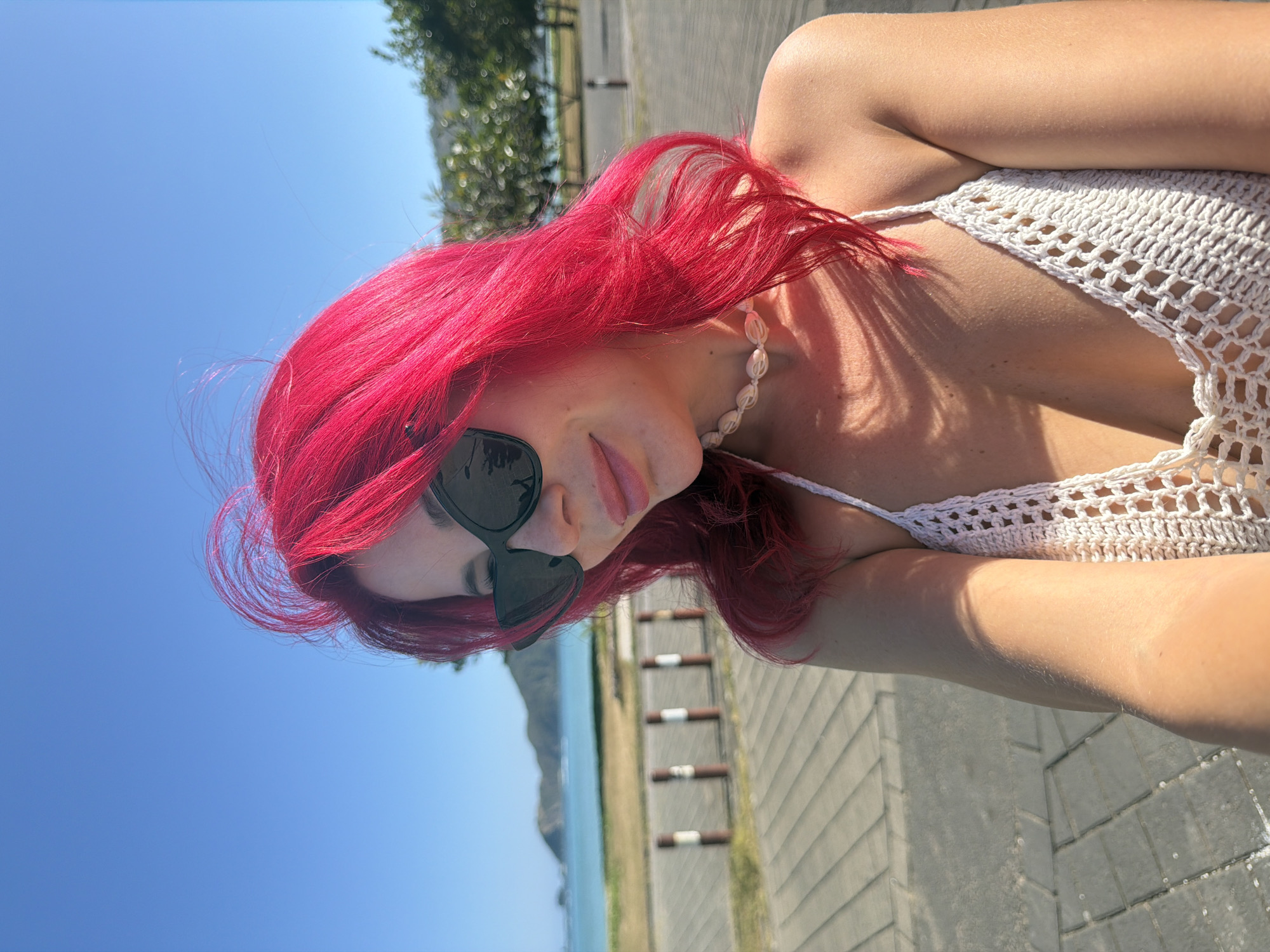 A woman with bright pink hair and sunglasses stands on a paved walkway near a beach. She is wearing a white crocheted bikini top and a shell necklace. In the background, there is the ocean, a sandy area, trees, and a distant hillside. The sky is clear and blue. The location is Kuta, Indonesia.