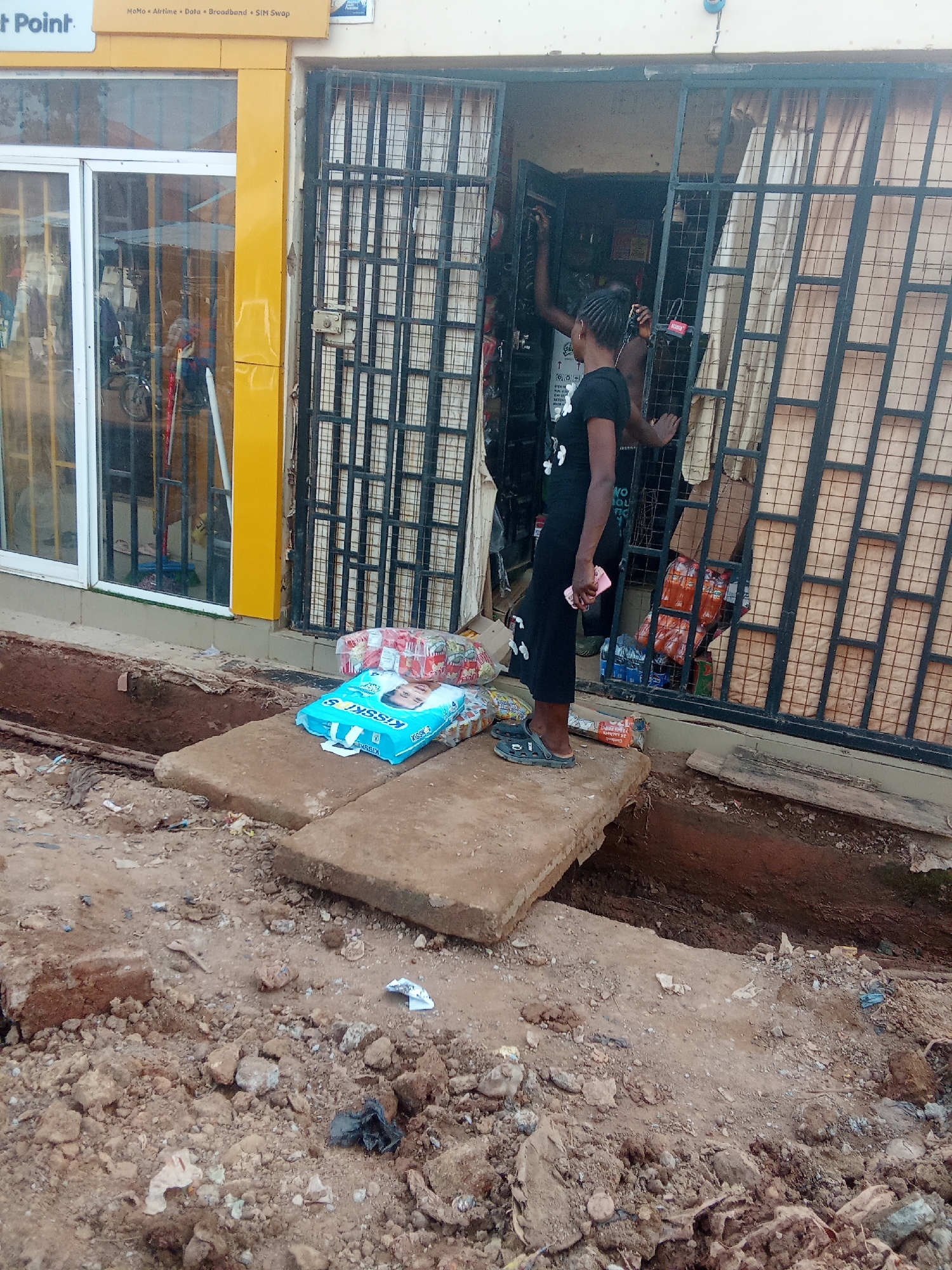 In Kagini, Nigeria, a street-level view captures a scene of construction and commerce. A narrow trench has been dug along the roadside, with concrete slabs laid across it to facilitate pedestrian movement. On these slabs, a package of "Kisskies" diapers and other bagged merchandise are placed.
Adjacent to the trench is a storefront with a bright yellow pillar and large glass windows. Inside, behind a black metal gate, shelves are stocked with various goods, including bottled drinks. Two individuals are visible near the entrance of the store. One person, wearing a black t-shirt and black pants, is standing with their back to the camera, holding a pink mobile phone. The other person, only partially visible inside the store, appears to be attending to the merchandise. The ground is a mixture of dirt, gravel, and debris, indicating ongoing work or neglect.