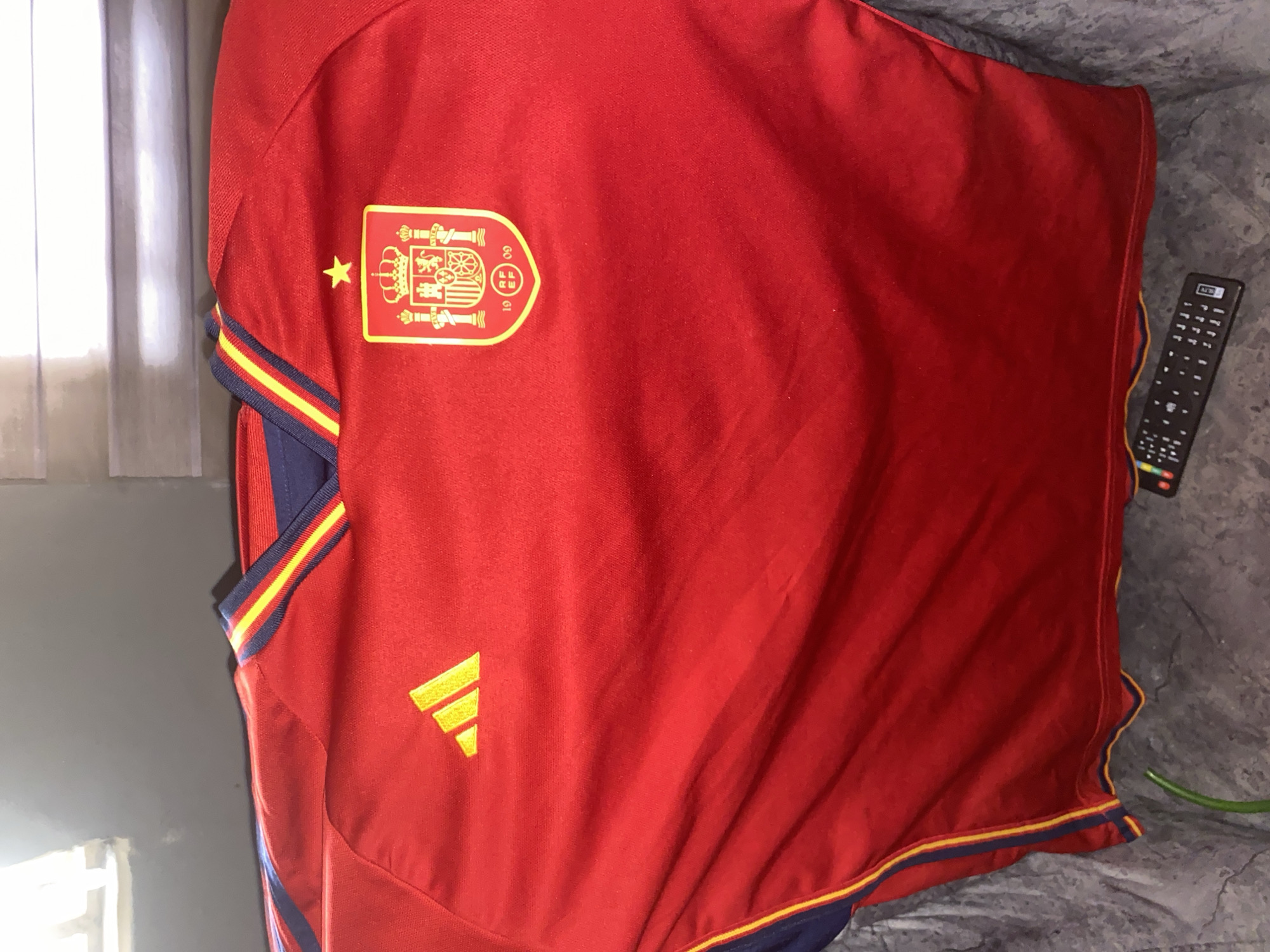 A red Spain soccer jersey is laid out on a gray surface, likely a bed or couch. The jersey has the Spanish national team badge on the left chest, a yellow Adidas logo lower down on the left, and yellow and blue trim on the sleeves and bottom hem. A black television remote control is visible on the right side of the image. There are no people visible. The setting appears to be indoors, possibly a bedroom or living room. The location provided, Hanwa, Nigeria, is not visually confirmed by the image.