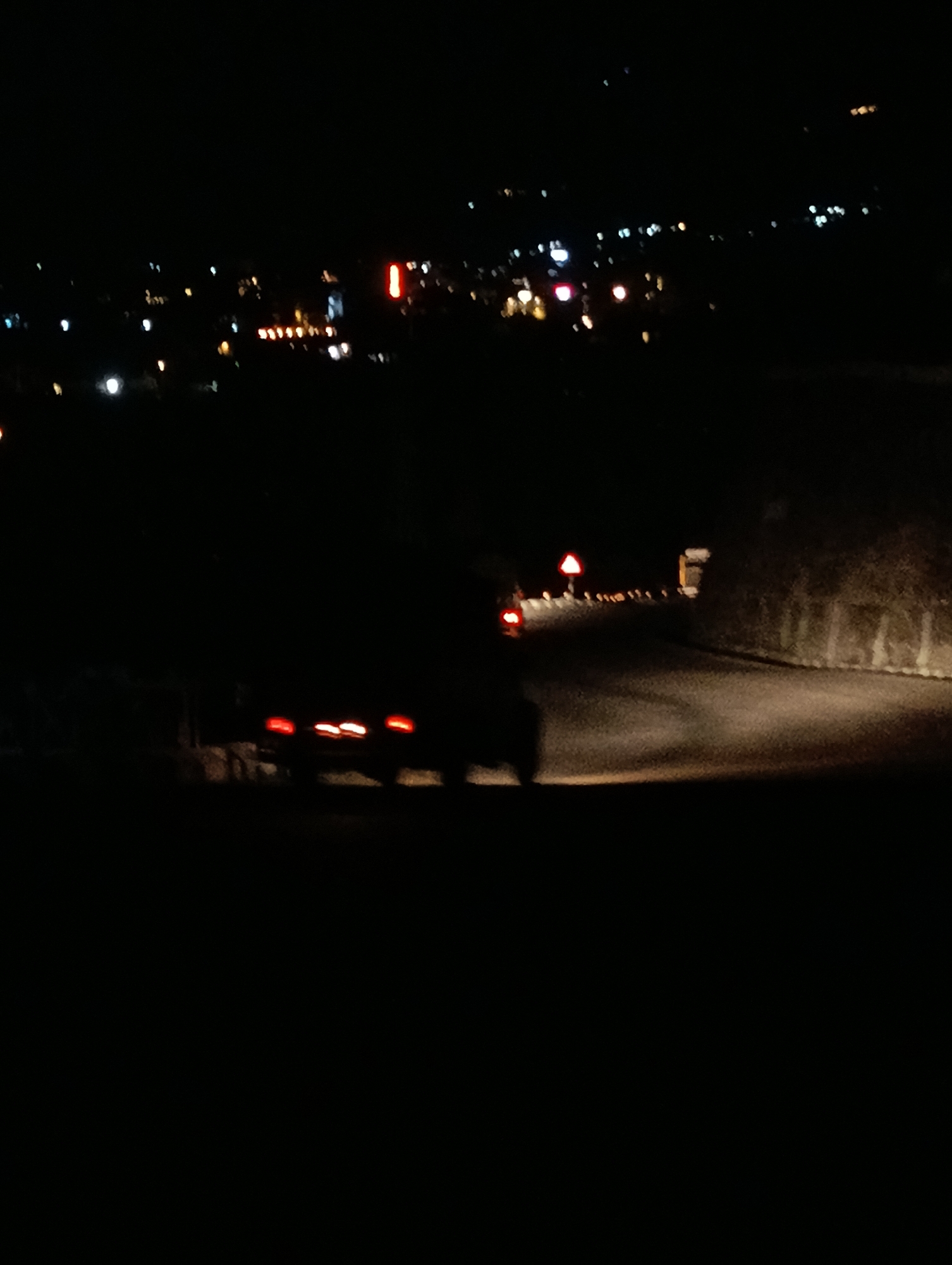 In the city of Kulang, India, at night, a dark, blurry scene unfolds. The image captures a road winding through a dark landscape, illuminated by the faint glow of distant lights and the red taillights of a vehicle. The road itself is visible as a lighter gray path, with the surrounding environment shrouded in darkness. Trees and vegetation appear as indistinct shapes along the right side of the road. A triangular warning sign with a red border is present on the left side of the road. The overall impression is one of low visibility and a sense of quiet or isolation, typical of a rural or suburban area at night.
