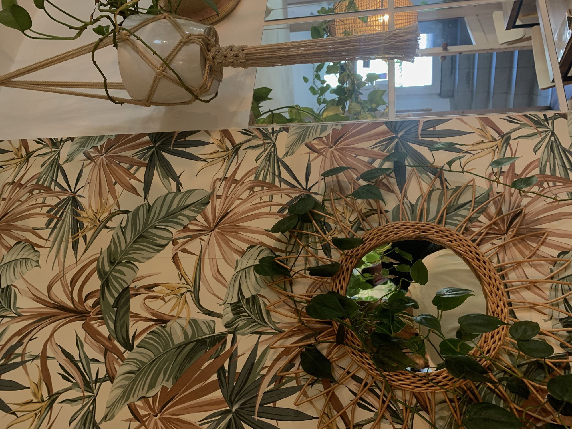The image depicts an indoor scene with a tropical-themed wallpaper. Prominently featured is a rattan-framed mirror with greenery draped over it. To the right, a white ceramic pot with a plant hangs in a macrame hanger. A reflection in a mirror shows a person wearing a light-colored shirt, and beyond that, a doorway with stairs leading up. This scene is located in Poznan, Poland.
