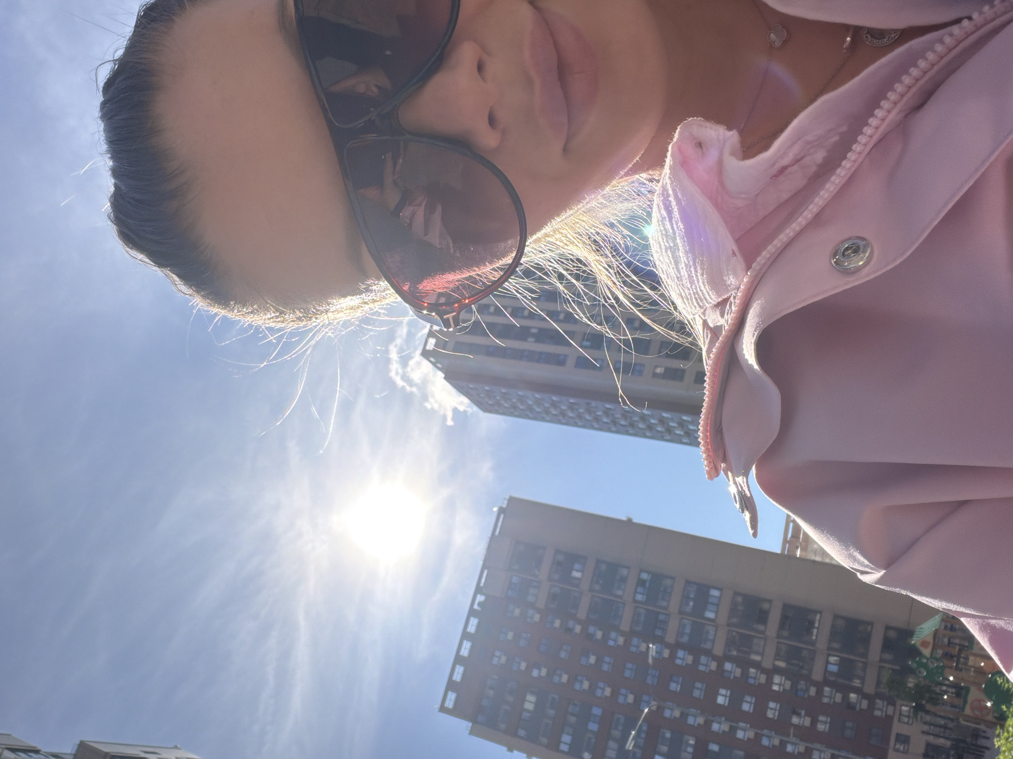 A close-up, low-angle selfie shows a woman from the chest up, looking into the camera. She is wearing large, dark-framed sunglasses, and her dark hair is pulled back. Her lips are painted a light pink. She is wearing a pink jacket, zipped partially up. The background shows a bright, sunny sky with some wispy clouds. Two tall, modern buildings are visible in the background, one to the left and one to the right. The building on the left is predominantly a light brown color with many windows, while the building on the right is darker and appears to be made of glass. The overall impression is a sunny day in a city. The location is likely Moscow, Russia, given the architecture of the buildings.