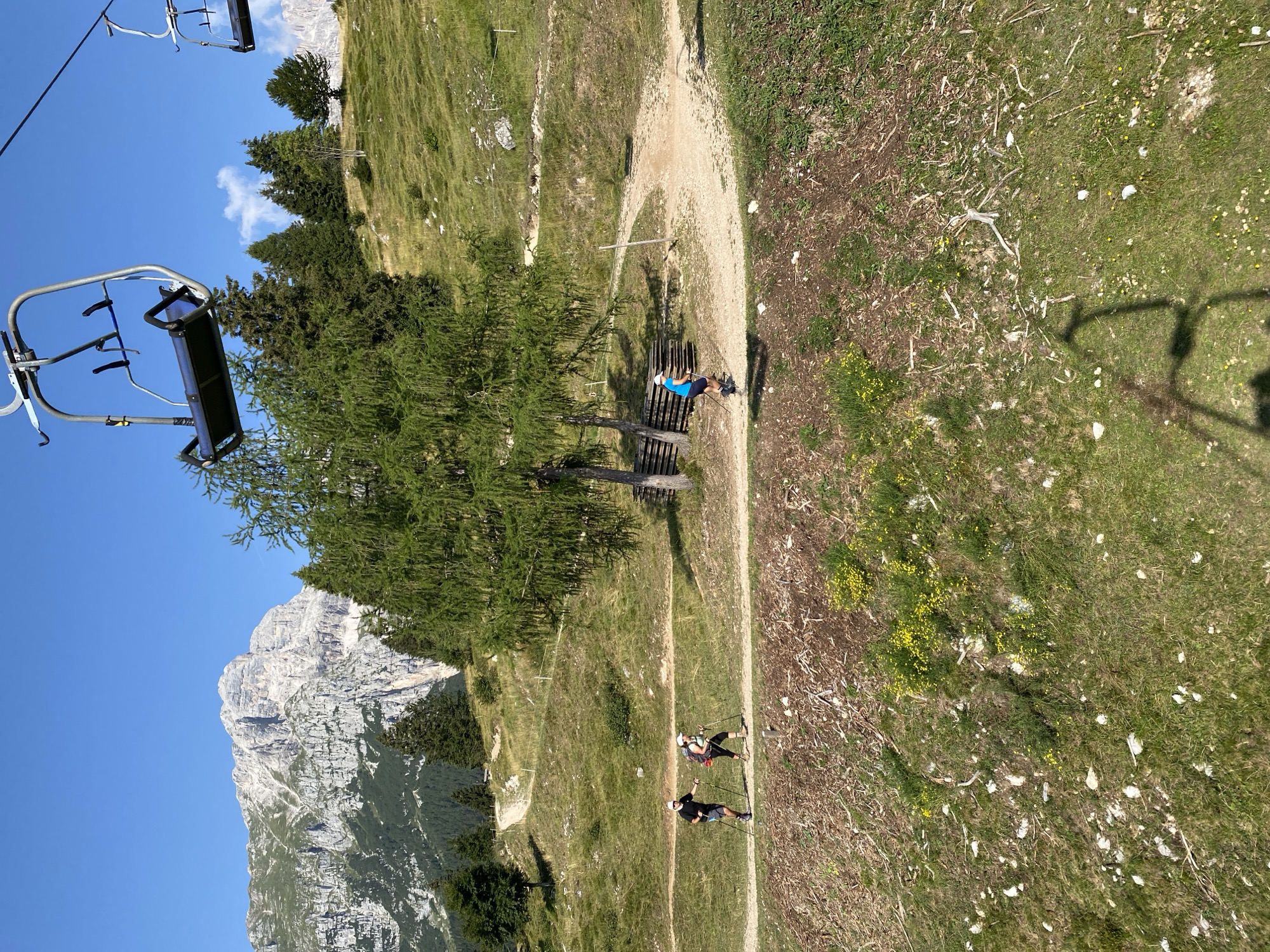 An outdoor shot on a sunny day shows a mountainous landscape with a clear blue sky. In the foreground, a ski lift is visible, with two empty chairs hanging. A dirt path winds through a grassy, sloped terrain dotted with trees. Three individuals are walking on the path. The first two, a man and a woman, are walking side-by-side, both wearing hats and carrying trekking poles. The man is wearing a black t-shirt and shorts, while the woman is wearing a tank top and shorts. A third person, wearing a blue t-shirt and shorts, is walking behind them. In the background, a large, rocky mountain range dominates the horizon, with dense green trees covering its lower slopes. The scene suggests a hiking activity in a mountainous region, possibly in the vicinity of Molveno, Italy, given the presence of ski lifts and similar mountain scenery.