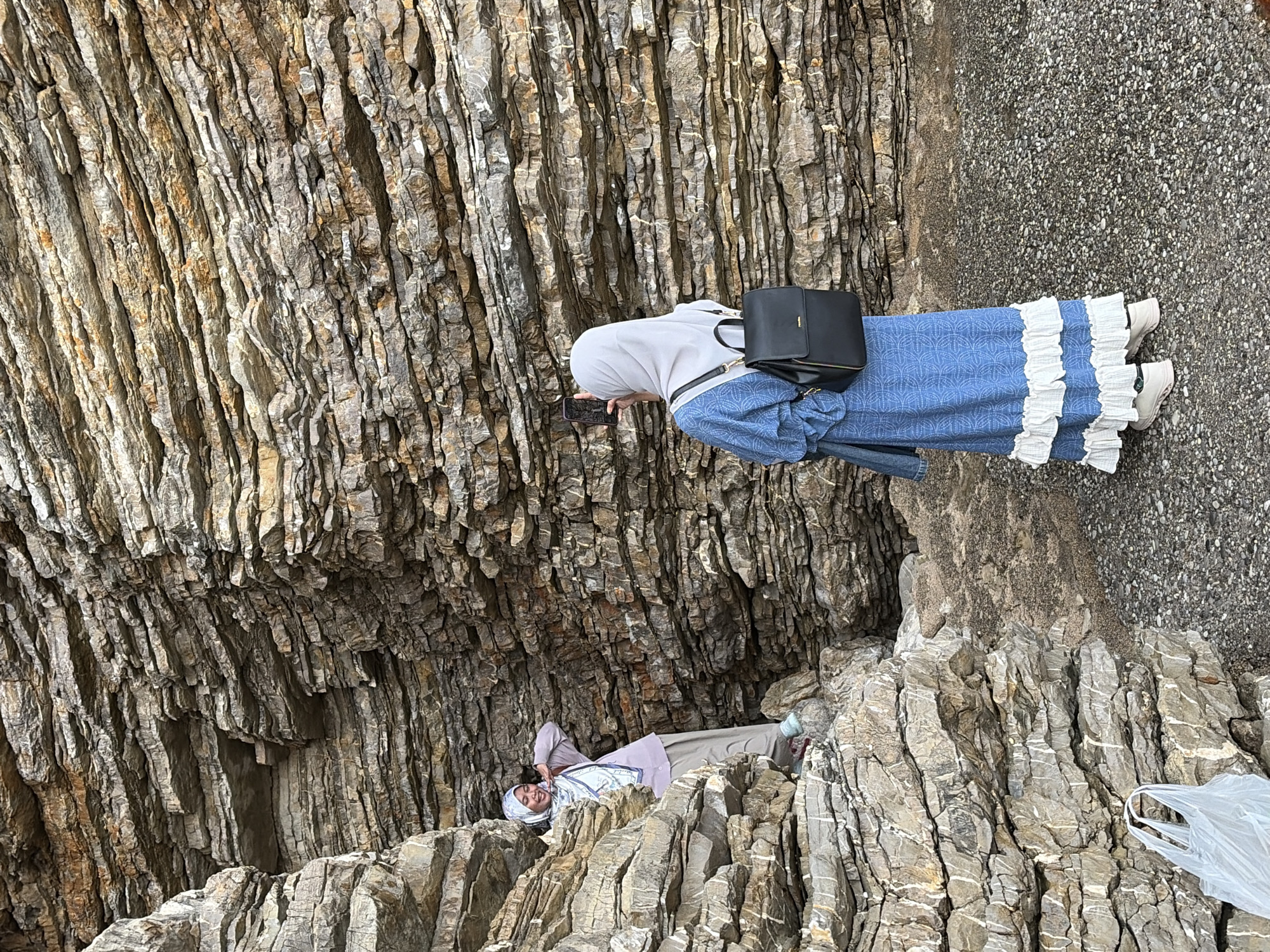 The image shows two people on a rocky shoreline or cliff in Budva, Montenegro. One person stands near a rock face, wearing a white hijab, a blue dress with white ruffles, a black shoulder bag, and white shoes. They appear to be holding and looking at a cell phone. The second person is lying amongst the rocks. They are wearing a light-colored hijab and a light-colored outfit. There is a white plastic bag among the rocks. The environment consists of stratified rocks and a paved area.