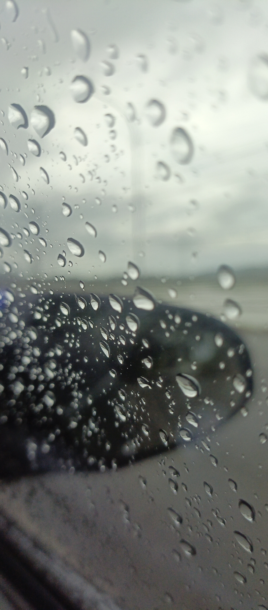 The image presents a close-up, vertical shot of a window covered in numerous water droplets, likely from rain. The droplets vary in size and shape, clinging to the glass, with many exhibiting distinct highlights and reflections that give them a crystalline appearance. They are sharply in focus, creating a textured foreground.
Through the translucent, water-covered glass, the background appears heavily blurred and out of focus. A dark, indistinct shape, possibly a car's side mirror or part of the vehicle's body, is visible towards the lower middle and right side of the frame. Above and beyond this, a soft, grey, and white expanse suggests an overcast sky or a distant, muted landscape.
The scene depicts a rainy or post-rain moment, viewed from inside a vehicle. The lighting is diffused and soft, characteristic of an overcast day. The overall impression is one of quiet contemplation, observing the outside world through a veil of rain. The location context provided is Glee Gogo, Indonesia. There is no visible text in the image.