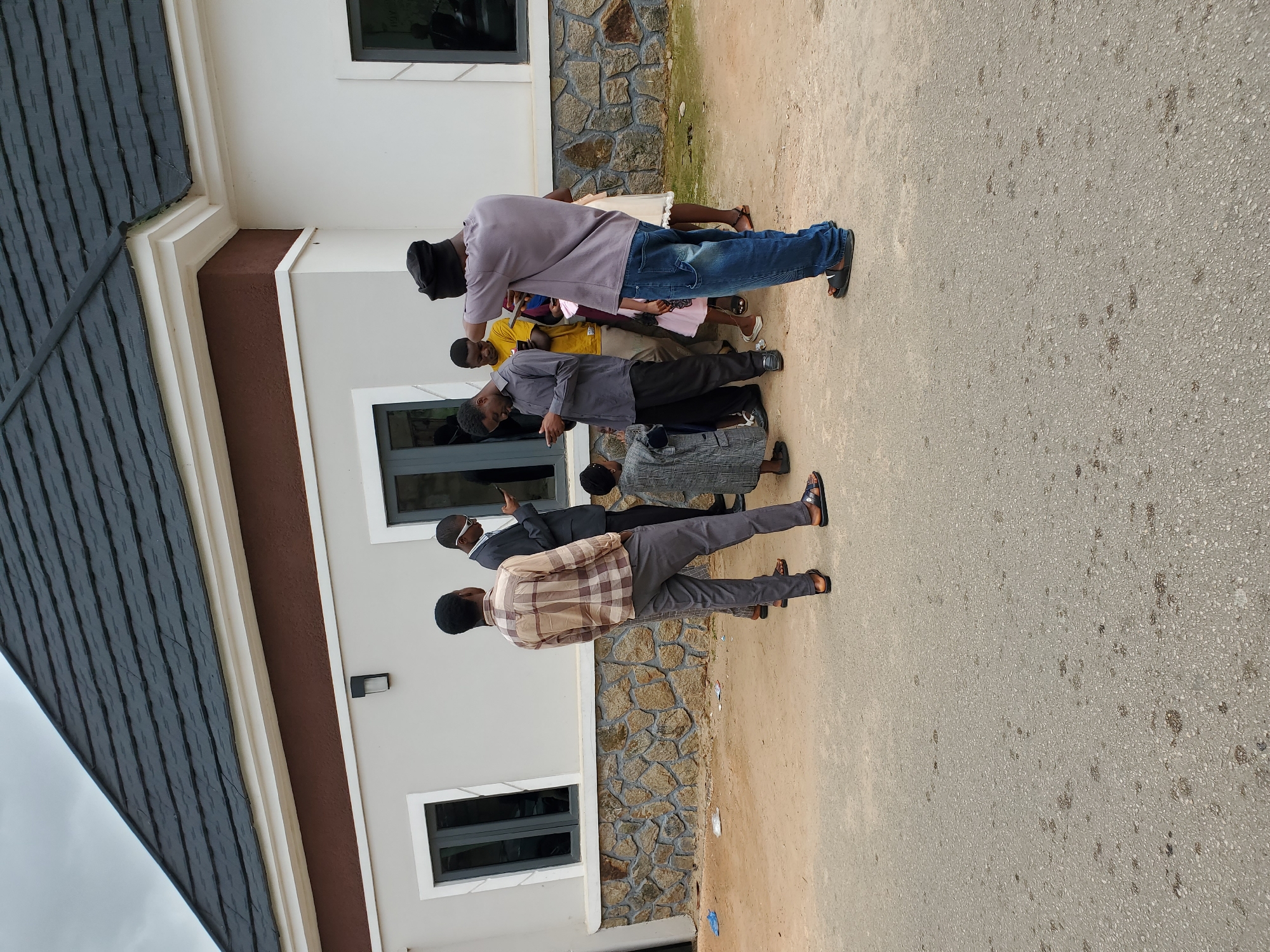 The image depicts a group of approximately seven people standing outside a building in what appears to be Jos, Nigeria. The building is a modern-style structure with a grey-tiled roof, white walls, and brown trim. It features stone accents and dark-framed windows. The people are of varying heights and dressed in casual attire, including button-down shirts, t-shirts, jeans, and pants. Some are wearing sandals, while others wear closed-toe shoes. One individual is holding a mobile phone. The group seems to be interacting or posing for a photo. The ground is paved with a grey asphalt surface.