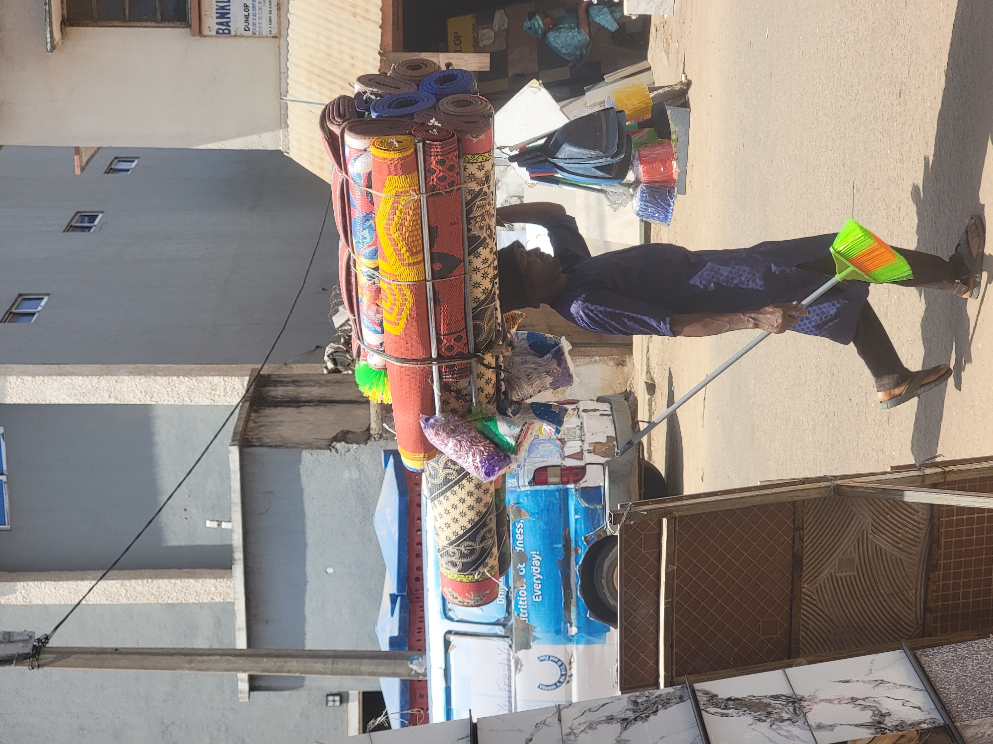 A man with dark skin stands on a paved street in Maiburiji, Nigeria. He is pulling a blue and white motor tricycle laden with rolled-up mats and other goods. The man is wearing a navy blue patterned top and shorts. He is holding a green and orange broom. Behind him, there is a tile display and various buildings. The sky is clear and sunny.