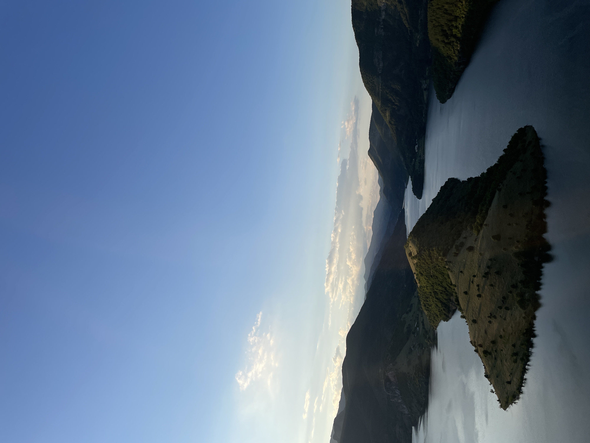 The image is an aerial view of a vast body of water surrounded by lush green mountains. The water, likely a reservoir or a large river, is calm and reflects the sky and surrounding landscape. A prominent feature is a long, narrow peninsula jutting out into the water, covered in sparse vegetation and a few scattered trees. The hills and mountains are densely forested in some areas, with rocky outcrops visible in others. The sky is a clear blue, with a few wispy clouds scattered towards the horizon, suggesting either early morning or late afternoon light. The lighting casts long shadows, highlighting the contours of the landscape. The scene is tranquil and natural, devoid of any visible human presence or man-made structures, with the exception of possibly a small building or two visible in the distance on the far bank of the water. This vista is characteristic of the area around Borovitsa, Bulgaria, known for its dramatic mountain scenery and reservoir lakes.