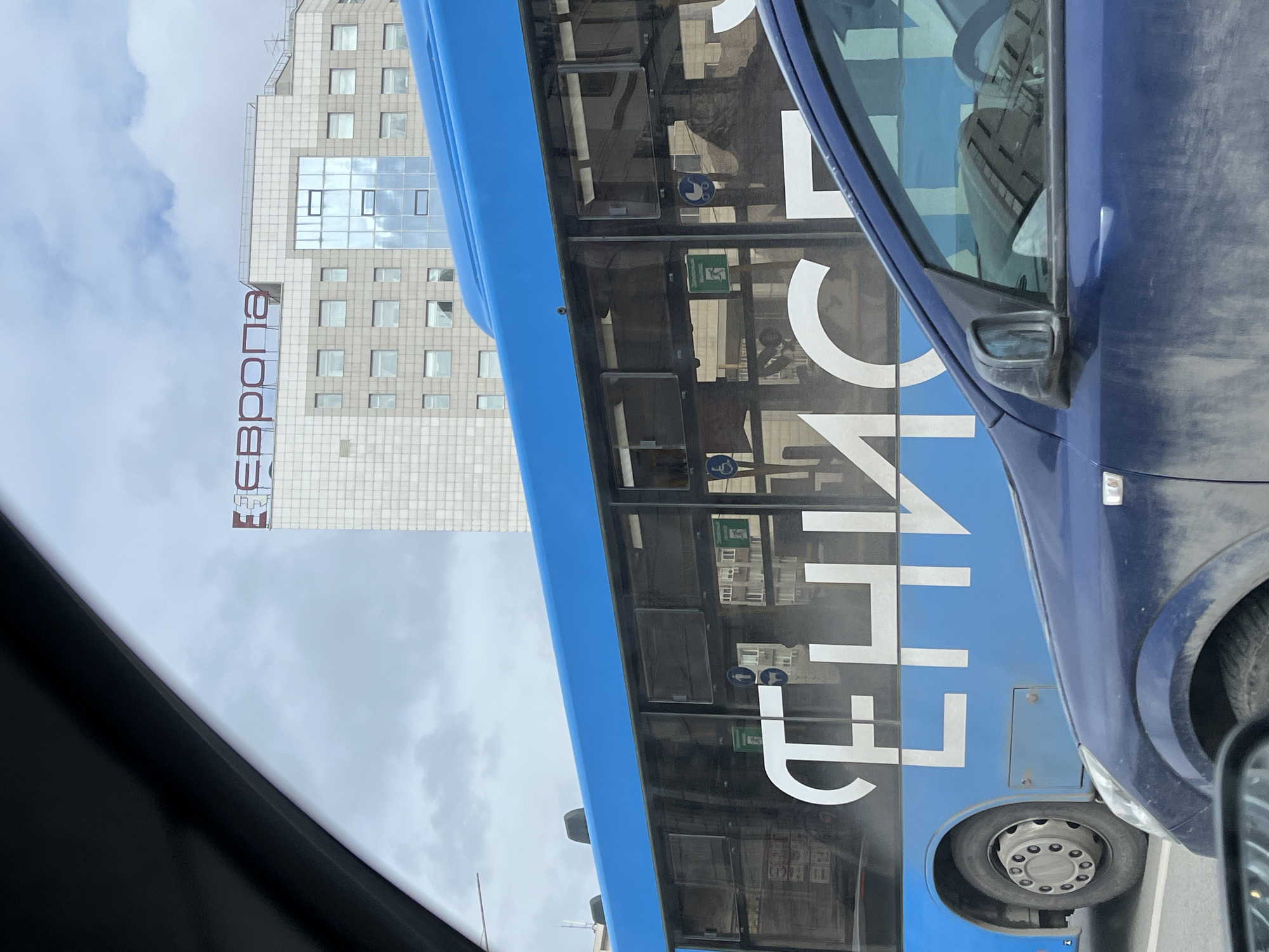 The image shows a view from inside a vehicle, with a blue bus adjacent to it. The word "СЕЛЬПО" is prominently displayed on the side of the bus in white, mirrored letters. Behind the bus, a tall, light-colored building is visible with the word "Европа" (Europe) written vertically on its side in a red-brown font. The sky is overcast, with clouds. The presence of the word "Европа" in Cyrillic suggests the location is in Russia. The "СЕЛЬПО" marking on the bus, a term commonly found in post-Soviet countries related to rural cooperative trade, further points to this region. The presence of both the "Европа" building and "СЕЛЬПО" bus suggests this picture was taken in Krasnoyarsk, Russia.