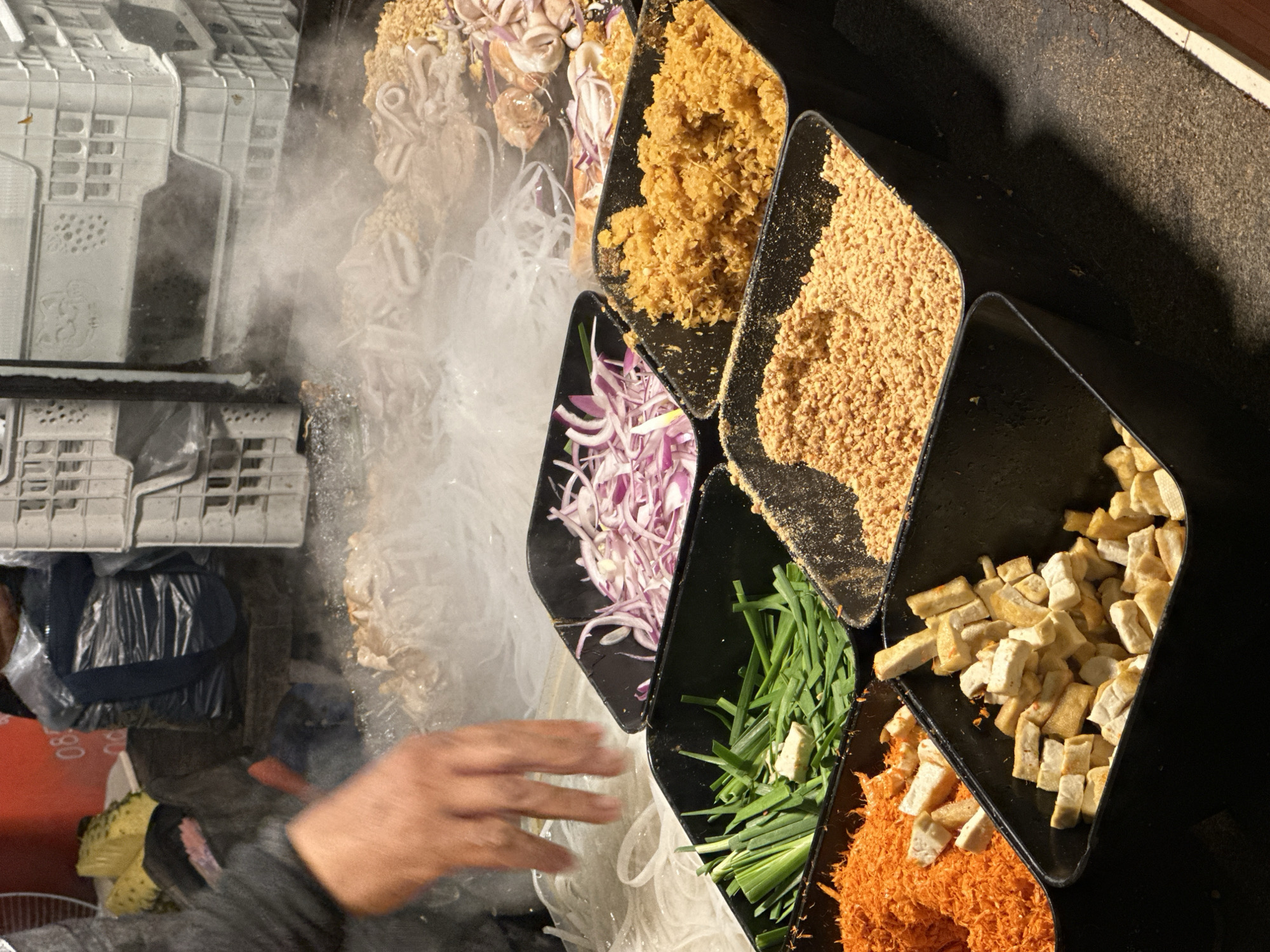 The image shows a street food vendor preparing Pad Thai in Pattaya City, Thailand. The vendor is arranging ingredients from separate black containers: bean sprouts, red onions, chives, orange shrimp floss, tofu, peanuts, and other fried ingredients. Steam rises from the bean sprouts. A hand reaches towards the bean sprouts and other ingredients. Shipping crates are visible in the background.