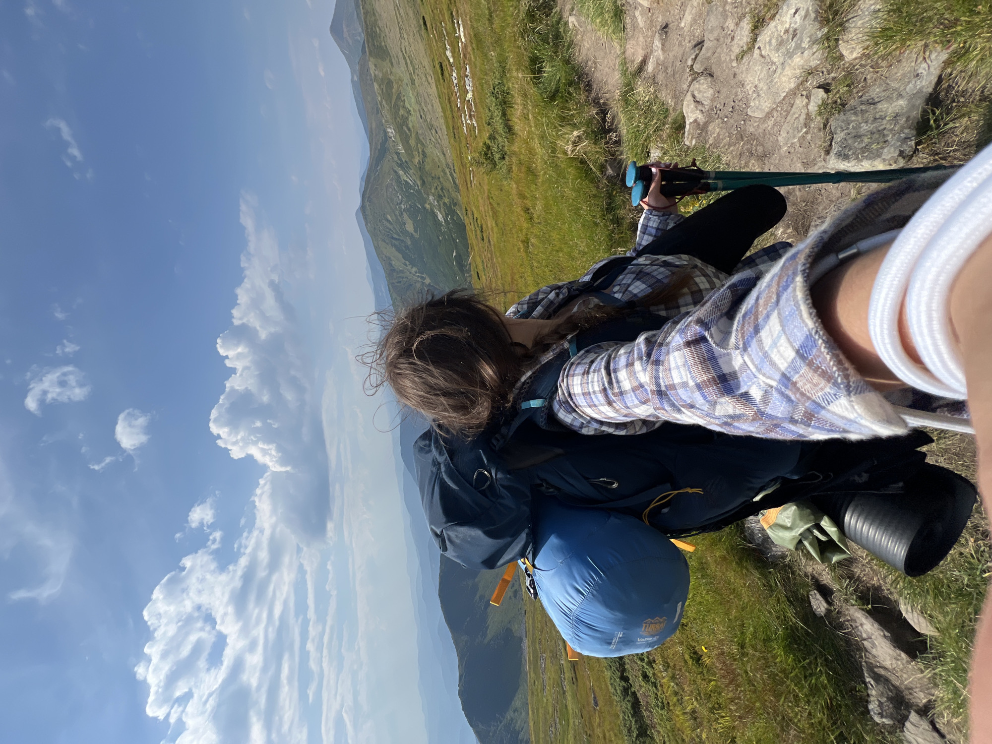 A first-person perspective shot captures a person hiking in the Carpathian Mountains near Luhy, Ukraine. The hiker, with long brown hair braided, is wearing a blue and white plaid shirt and black pants. They are carrying a large navy blue backpack, a blue sleeping bag attached to the side, and a black sleeping mat. In their right hand, they hold two hiking poles with blue grips. The background features rolling green mountains under a bright blue sky with scattered white clouds. A dirt path winds through the grassy, rocky terrain.