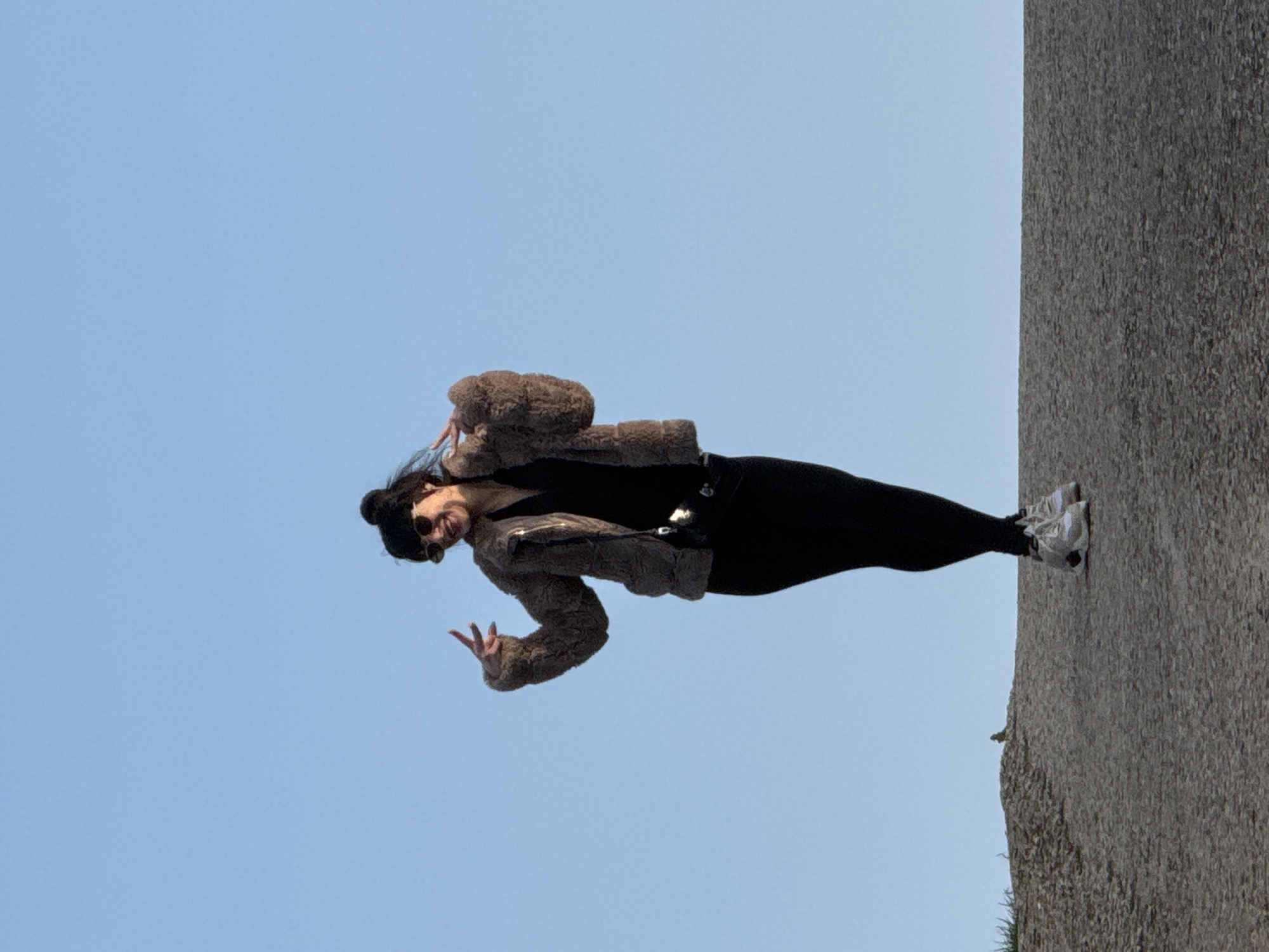 A woman wearing a brown jacket, black pants, and white sneakers stands on a textured grey stone structure against a clear blue sky. She wears sunglasses and has her hair in a bun. She is holding her right hand up in a peace sign. The location is Balgarevo, Bulgaria.