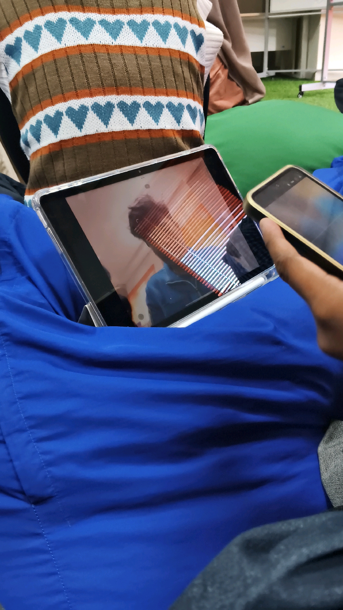 An individual is seated on a large, blue beanbag, positioned indoors. The person is wearing a knitted sweater vest with a pattern of alternating brown and white stripes, adorned with blue heart shapes. In their lap, they hold a tablet device, its screen reflecting a person in a blue garment and horizontal blinds. To the right of the tablet, another hand is visible holding a smartphone. The background suggests an indoor setting with green carpeting and shelving units. The scene takes place in the city of Lohbener, Indonesia.