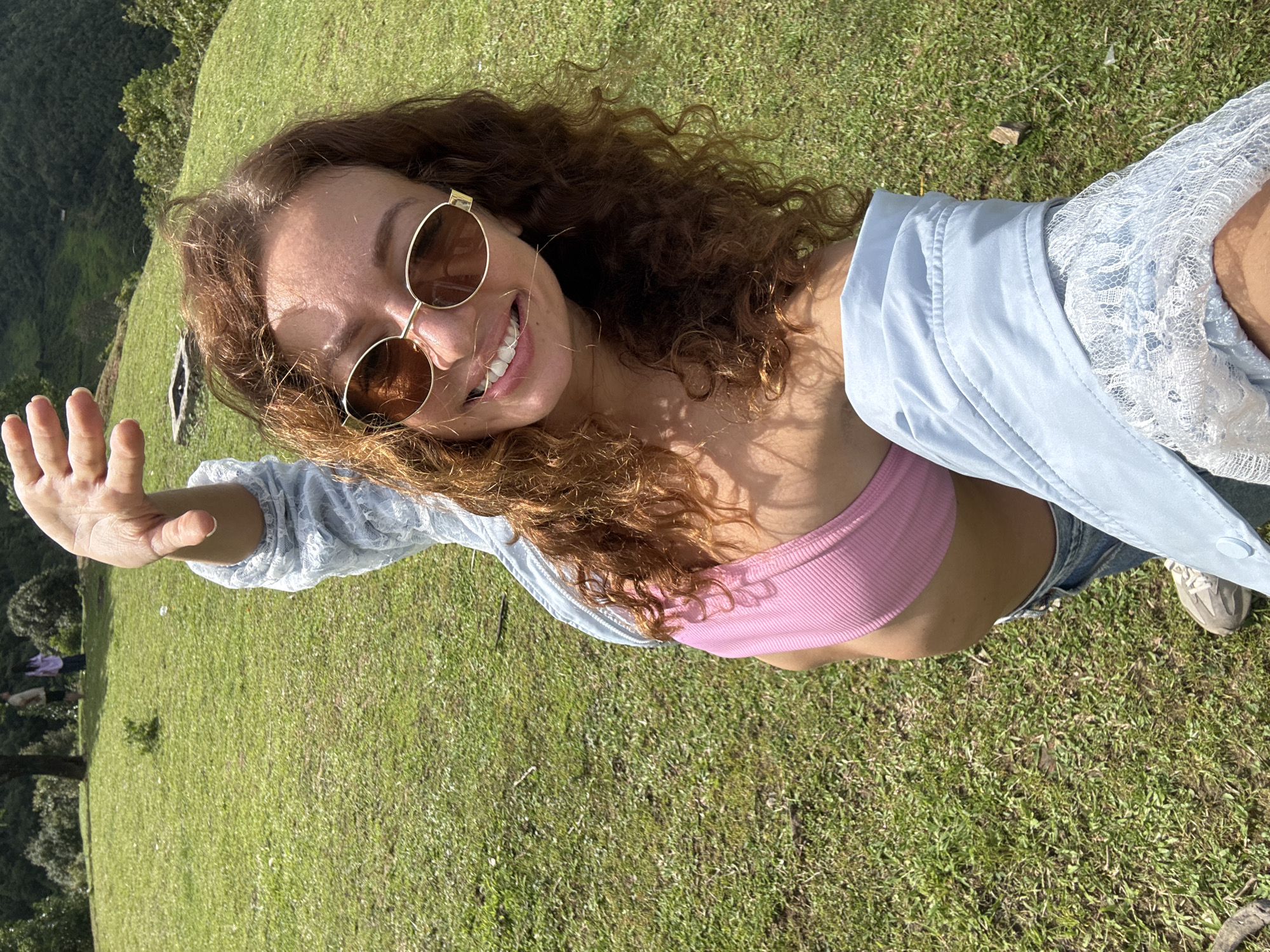 A young woman is depicted in a selfie taken on a sunny day in Tu Le, Vietnam. She is positioned in the foreground, smiling broadly with teeth visible, wearing gold-rimmed sunglasses, a pink ribbed crop top, and a light blue jacket featuring white lace detailing on the sleeve. Her midriff is exposed, and her left hand is raised towards the upper left corner of the frame. The background is a green grassy hillside. Two indistinct human figures are standing in the distant mid-ground. A square, light-colored concrete structure is embedded in the grass to the left of the woman's head. Dense green trees and hills occupy the far background. The scene is brightly illuminated by natural light.
