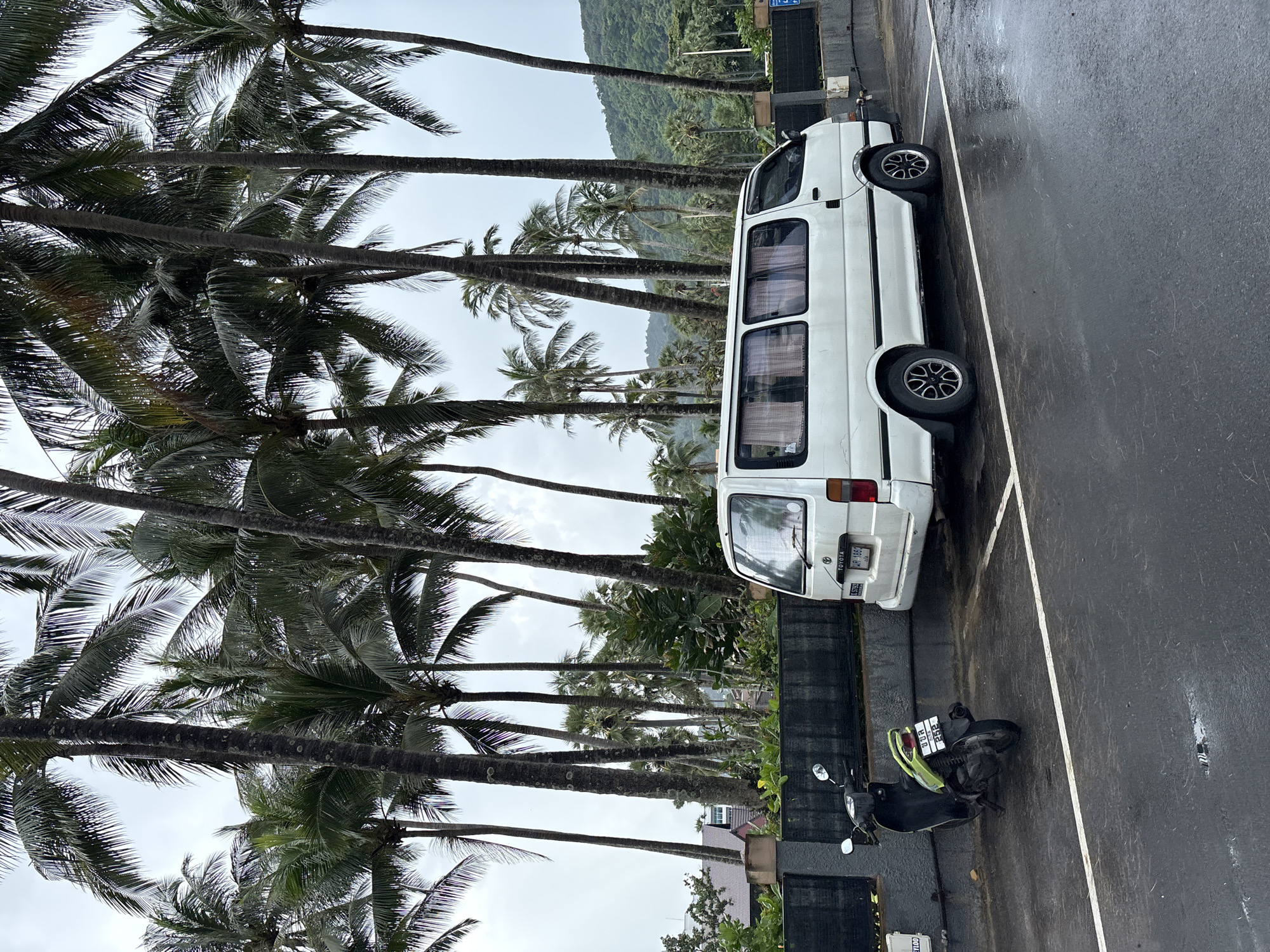 In Karon, Thailand, a white minivan is parked on a wet, asphalt surface. The minivan is positioned in a parking space marked by white lines. A green and black scooter is parked to the left of the minivan, also on the wet asphalt. Behind the vehicles, a dark gray wall runs horizontally, with a dense arrangement of tall palm trees towering above it. The sky is overcast, and a green hillside with more palm trees is visible in the background. The overall scene suggests a tropical environment, likely a parking area near a beach or resort, under a cloudy sky.